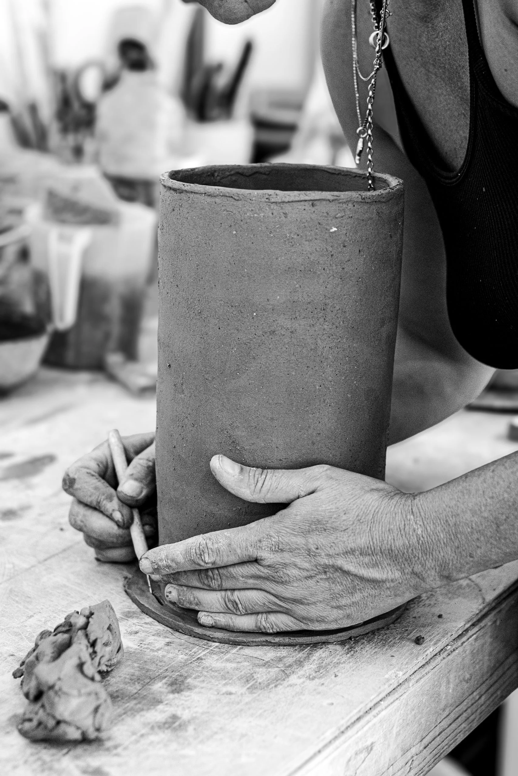 A person shaping a tall, cylindrical clay pot in a pottery studio, holding it with both hands, with workshop tools and materials in the background.