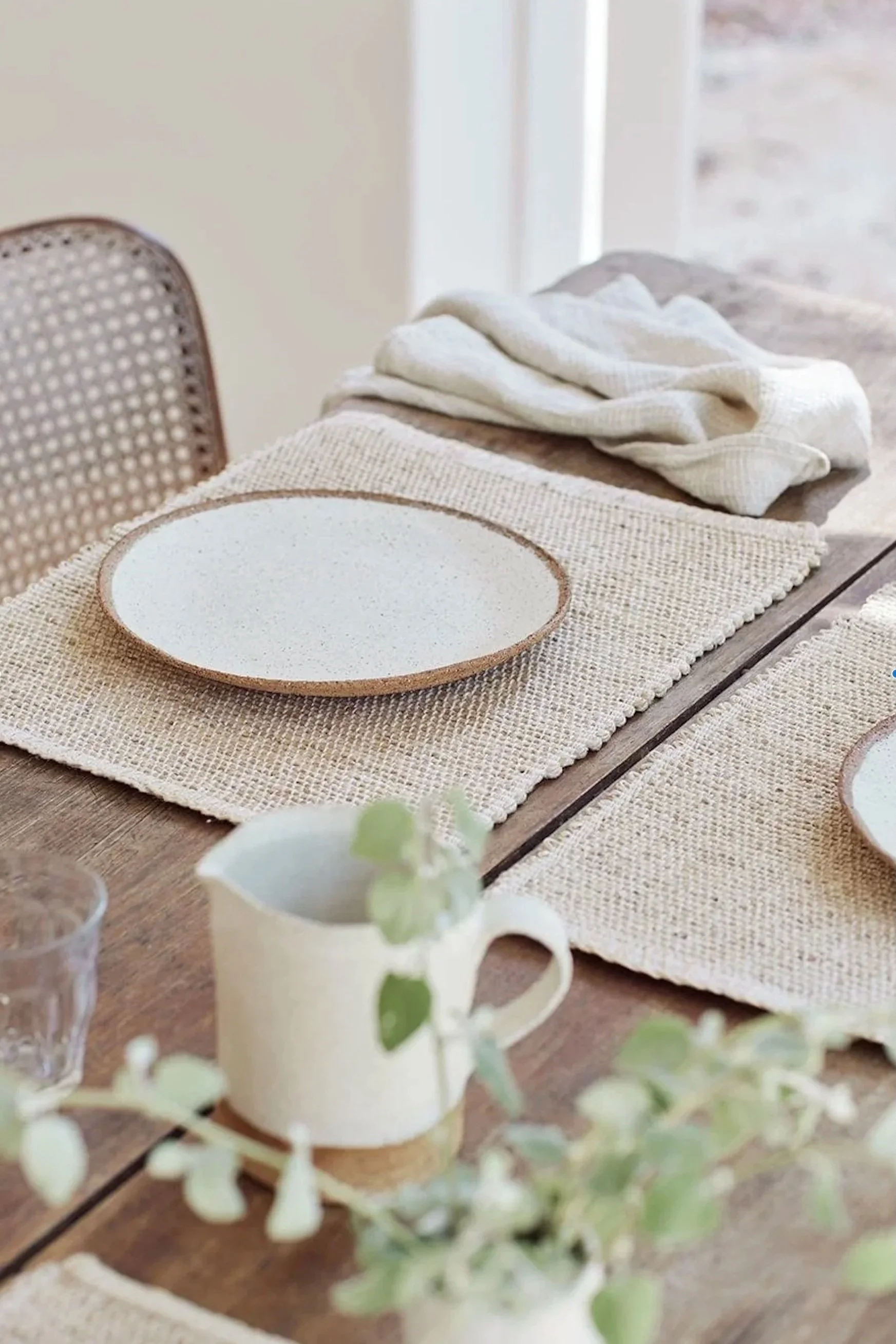 A dining table with beige woven placemats, a white ceramic bowl with a brown rim, a white mug with a sprig of greenery, and a white cloth napkin. The table and chairs are in a bright, cozy room with natural light.
