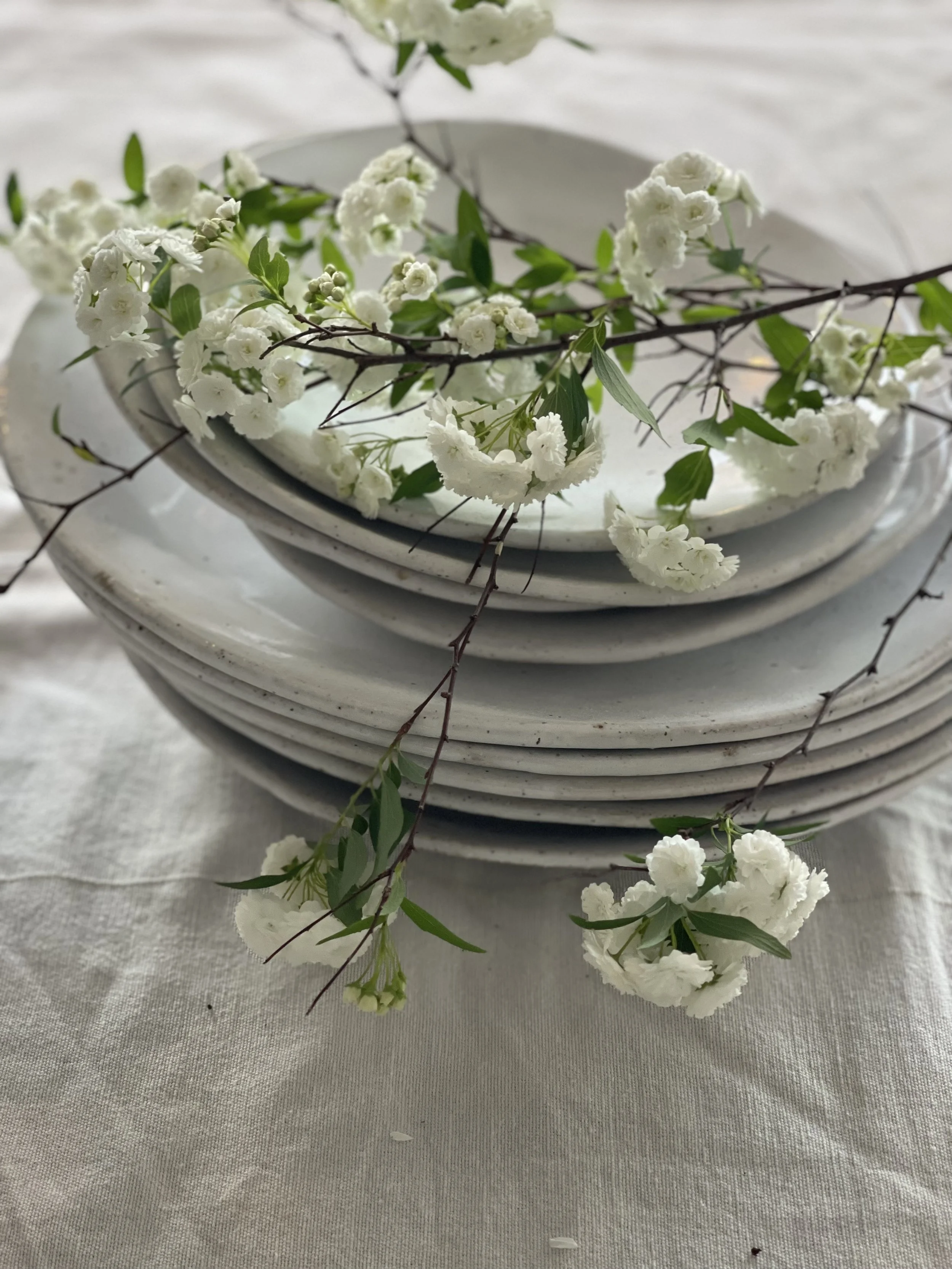 Stacked white plates with white flowers and green leaves on top, on a white tablecloth.