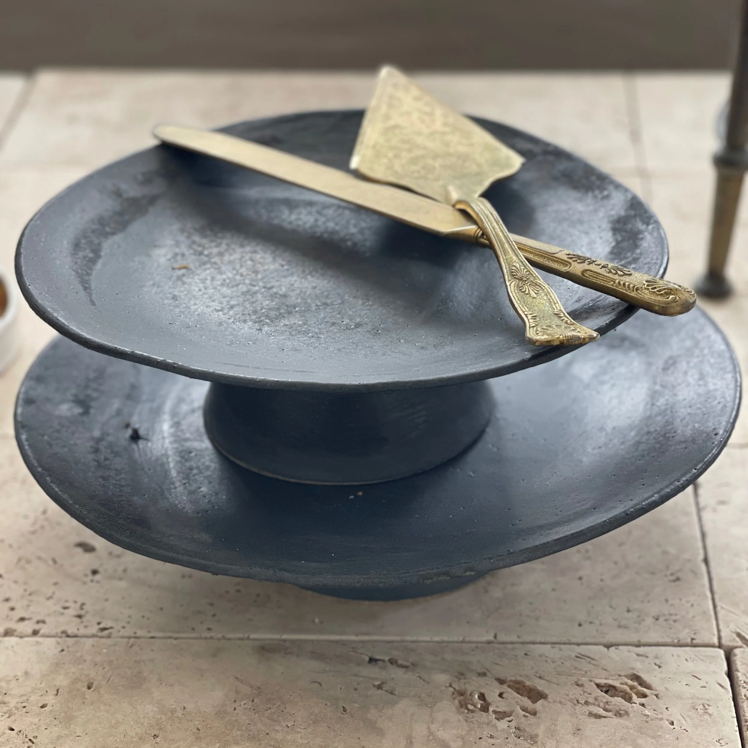 A black two-tiered serving stand with a gold knife and cheese slicer on top, placed on a tiled surface.
