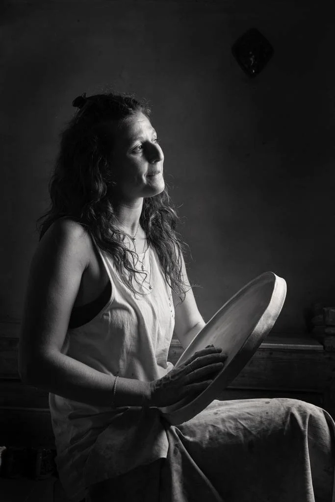 A woman sits on a bench holding a large round wooden tray, looking thoughtfully into the distance. The photo is in black and white.
