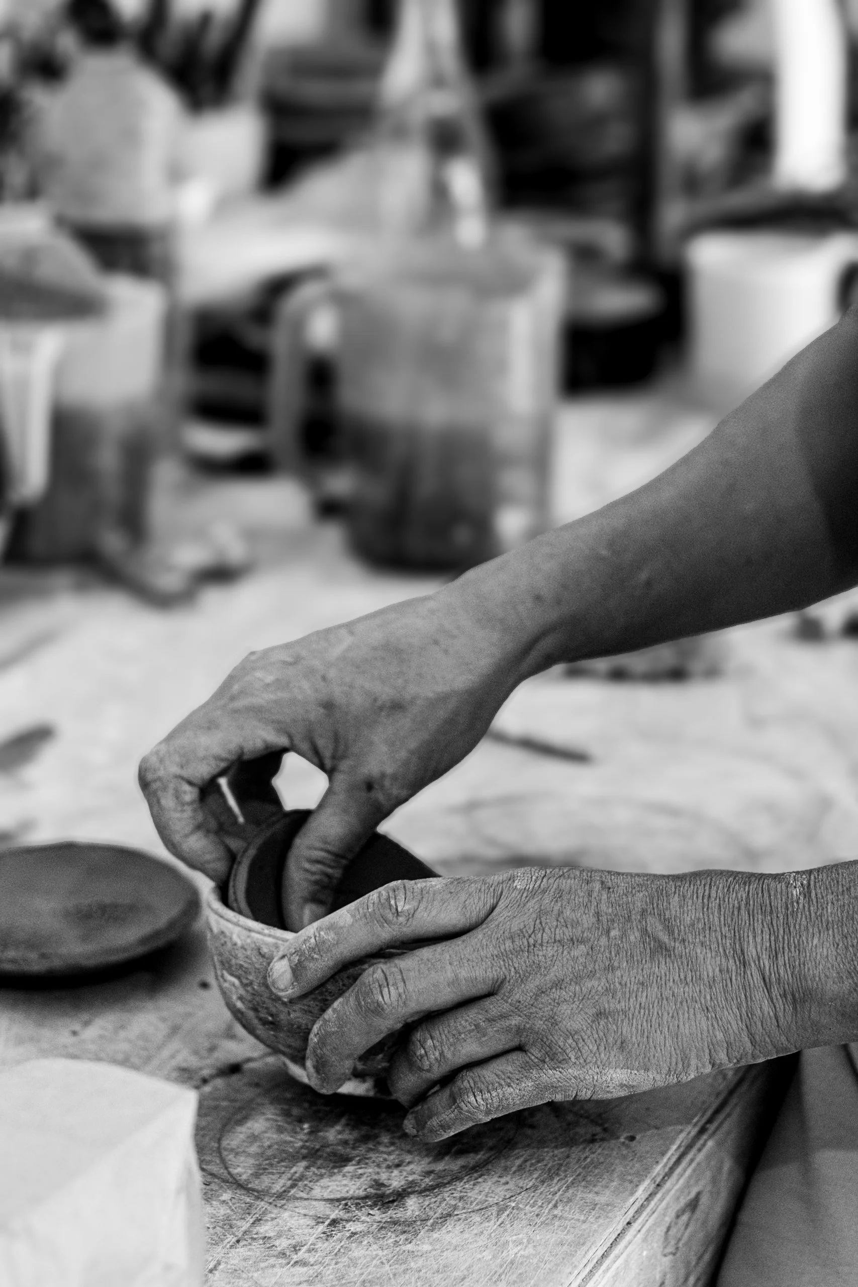 Close-up of hands working on a pottery piece in a workshop. The person is using their fingers to mold or smooth the pottery on a wooden table, with various pottery tools and equipment in the blurred background.
