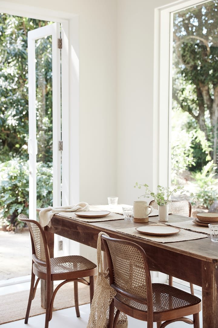 A dining area with a wooden table set with beige dishes, a white pitcher, and a small plant, near an open door and large window revealing greenery outdoors.