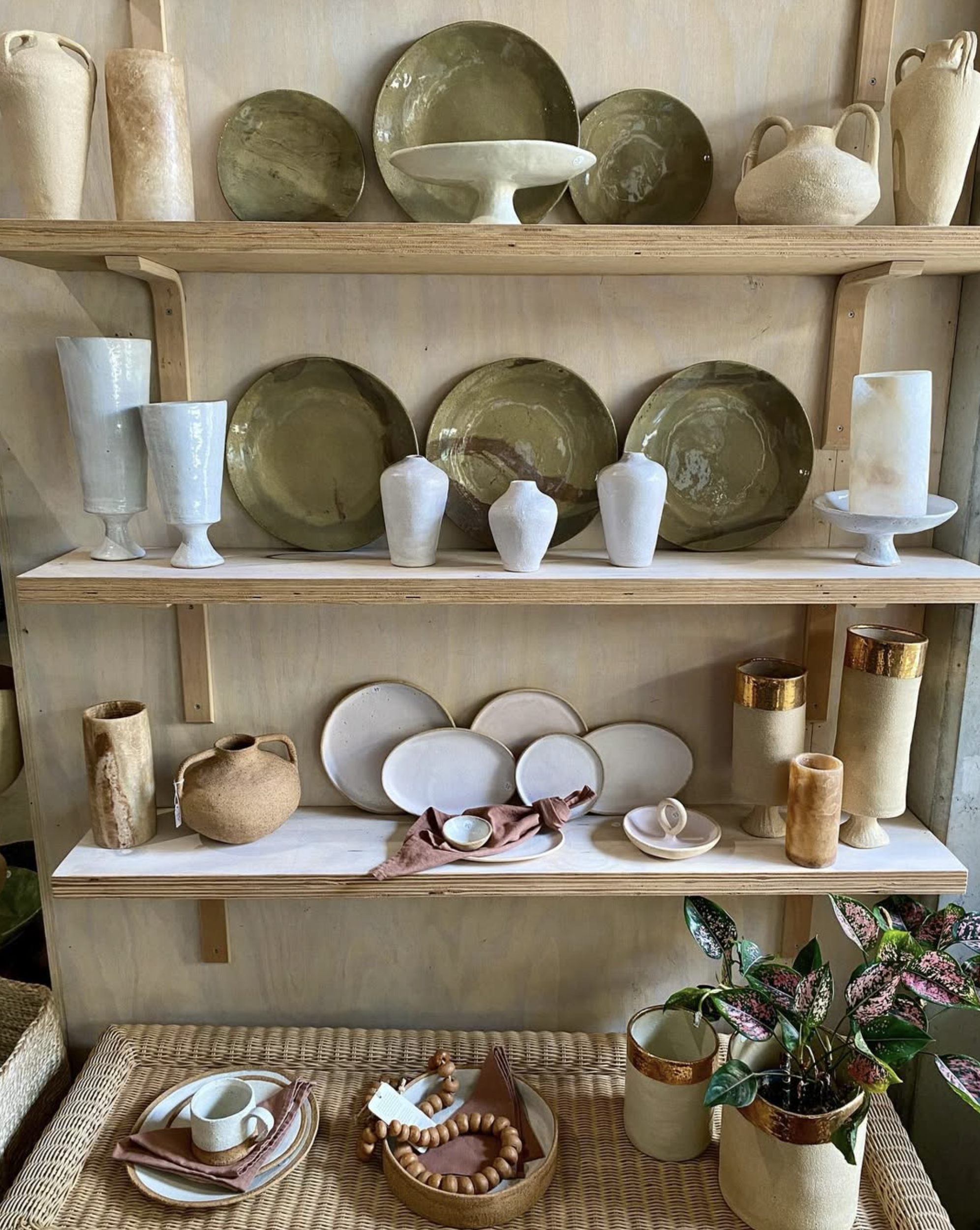 Display of decorative pottery and ceramics on wooden shelves and a woven table, including vases, plates, jars, and candles in neutral and earthy tones.