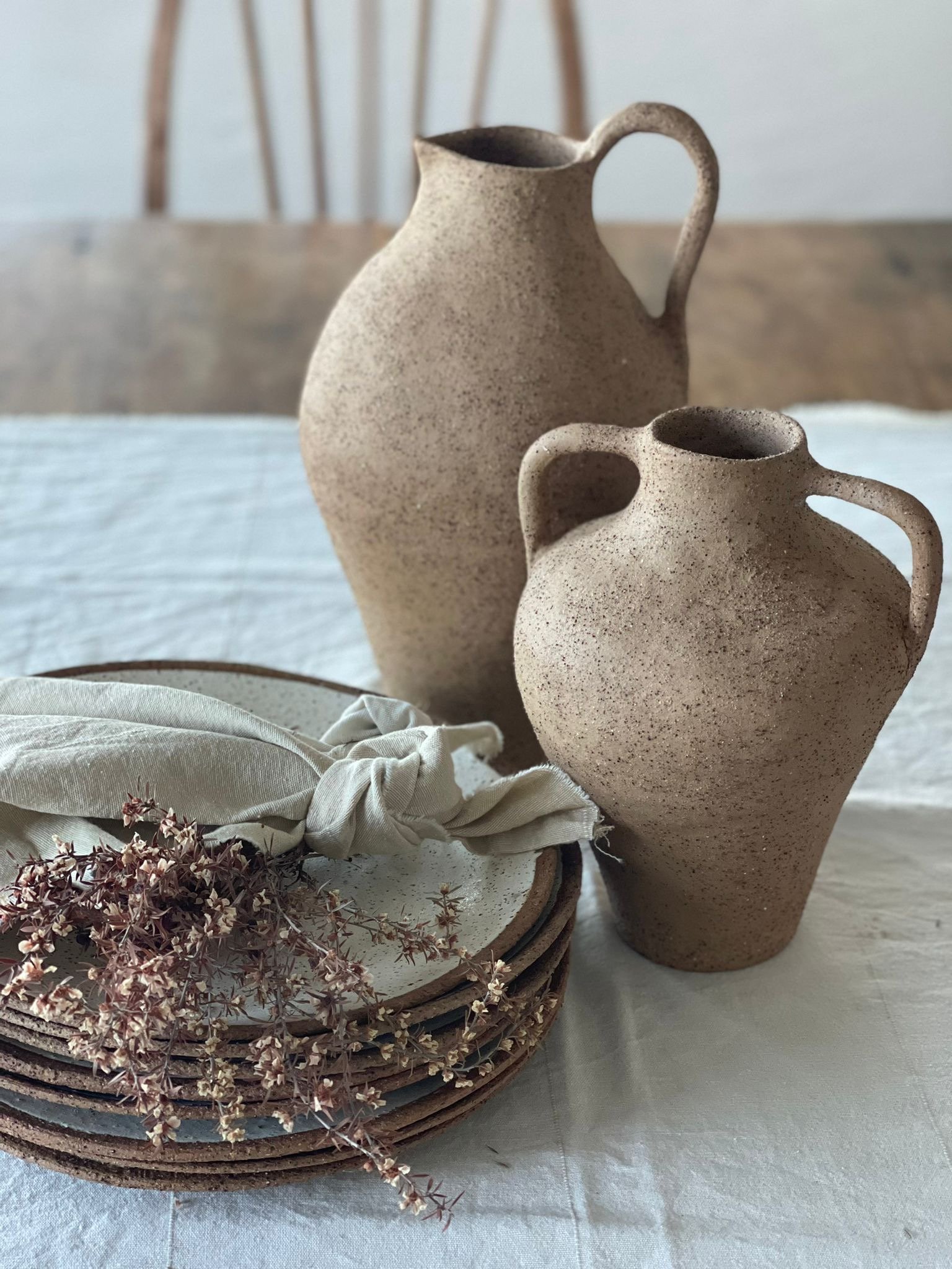 Two beige ceramic vases with handles, a stack of three ceramic plates with a cloth napkin and dried flowers on top, all arranged on a table with a white tablecloth.