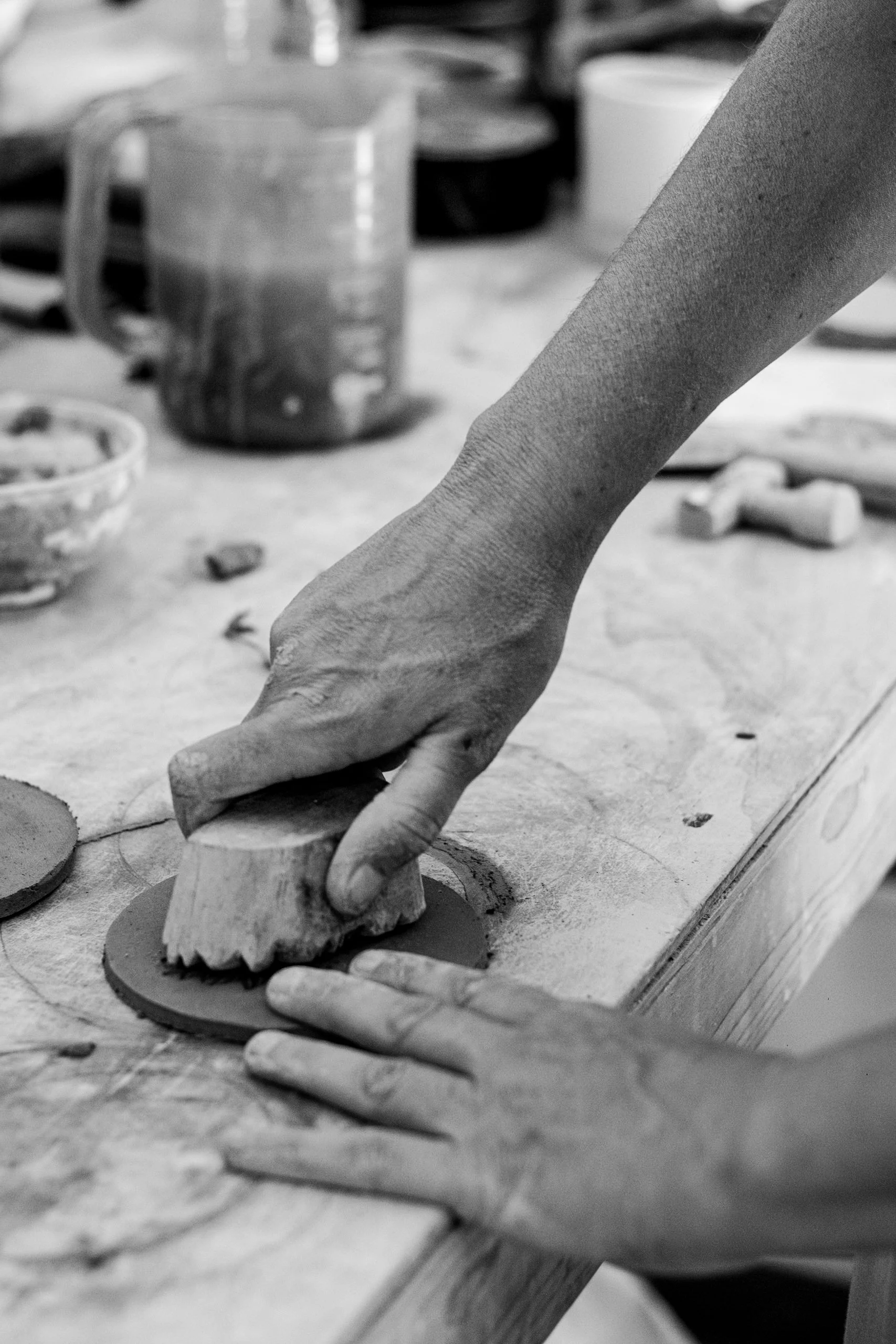 Close-up of a hand pressing clay with a wooden stamp on a workbench, with pottery tools and cups in the background in a pottery studio.
