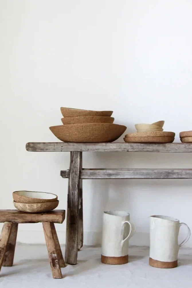 Stacked clay bowls on a rustic wooden shelf, with additional bowls on a small wooden stool, and two ceramic pitchers on the floor against a white wall.