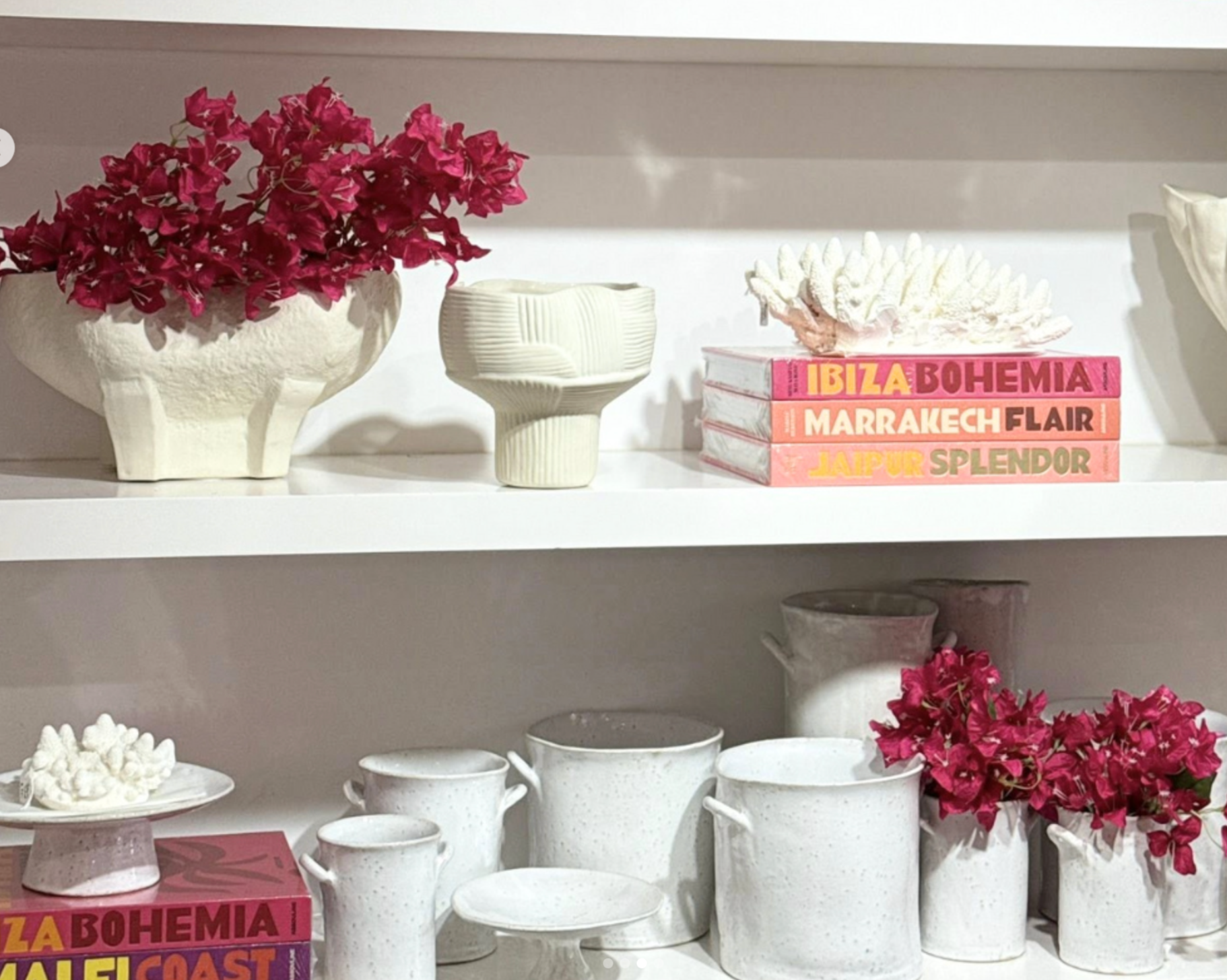 White shelves holding pink flowers in white vases, white ceramic cups and pitchers, decorative books, and coral sculpture.