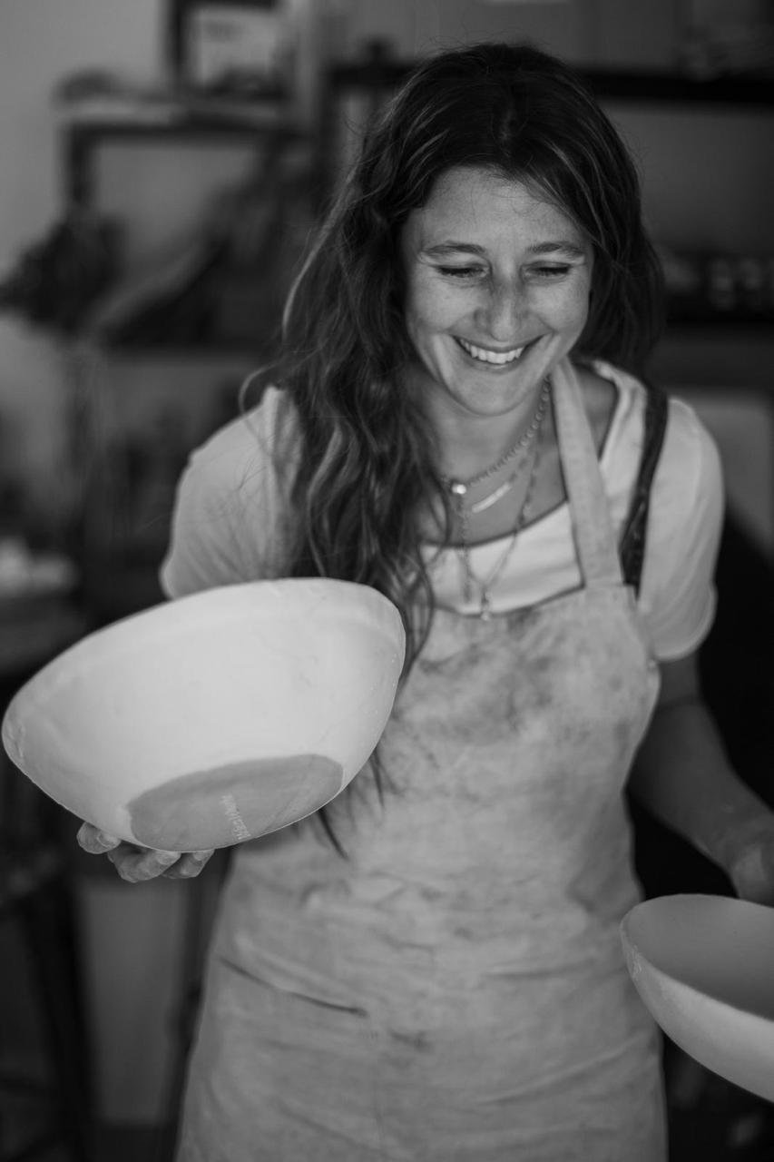 A woman with long dark hair, smiling, wearing an apron, holding a bowl, in a kitchen setting.