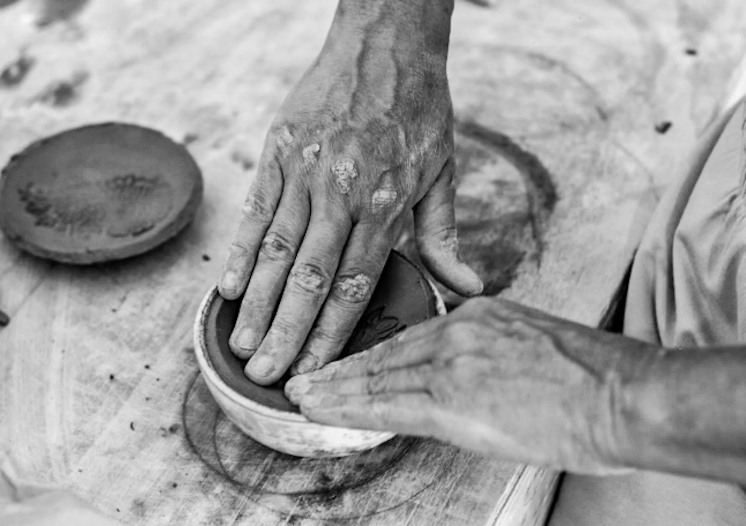 Hands shaping a clay bowl on a wooden surface, with two other clay bowls nearby, all appearing in black and white.