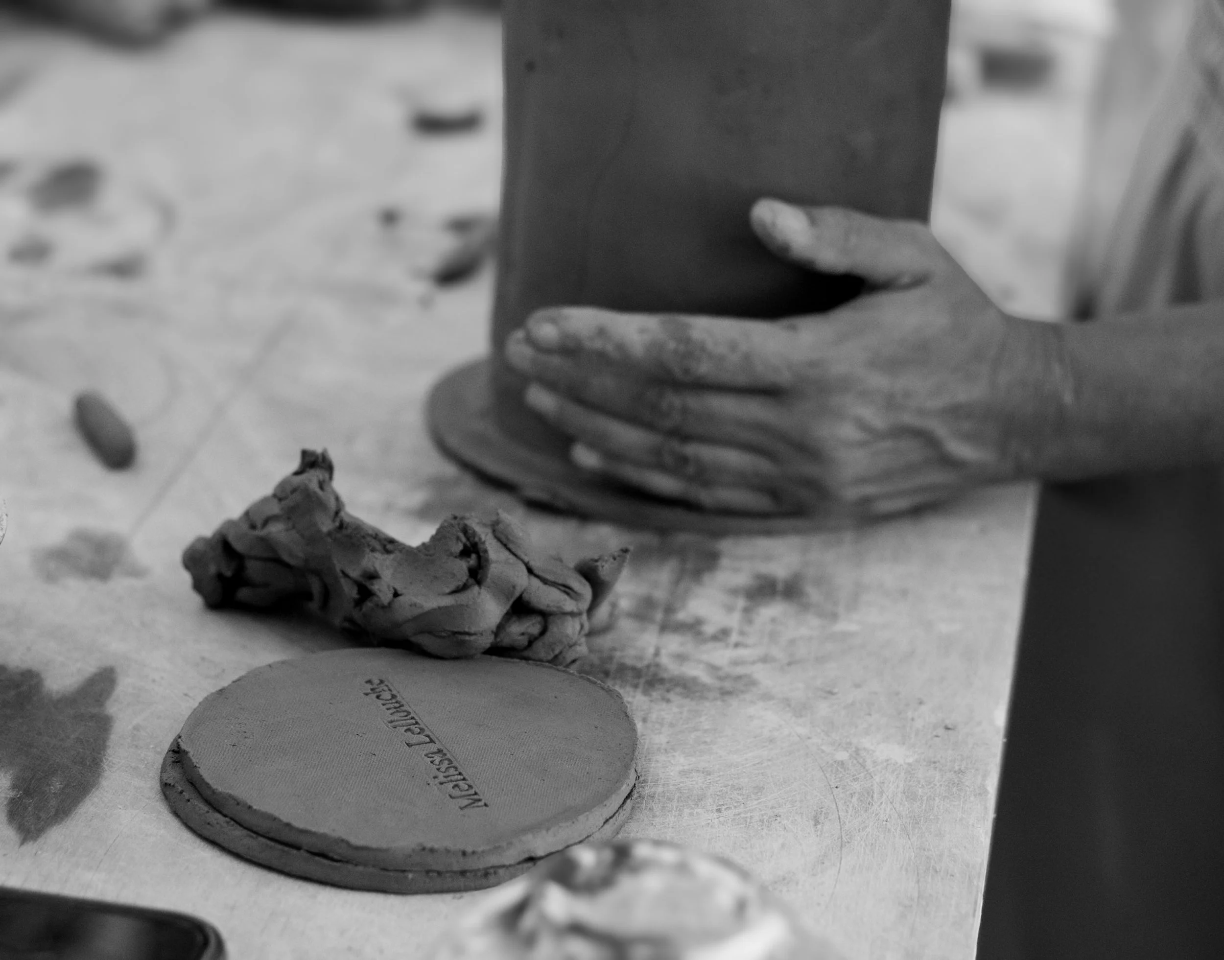 A person shaping clay with their hands in a pottery or sculpting workspace. There are clay tools and a clay-coated coaster on the table.