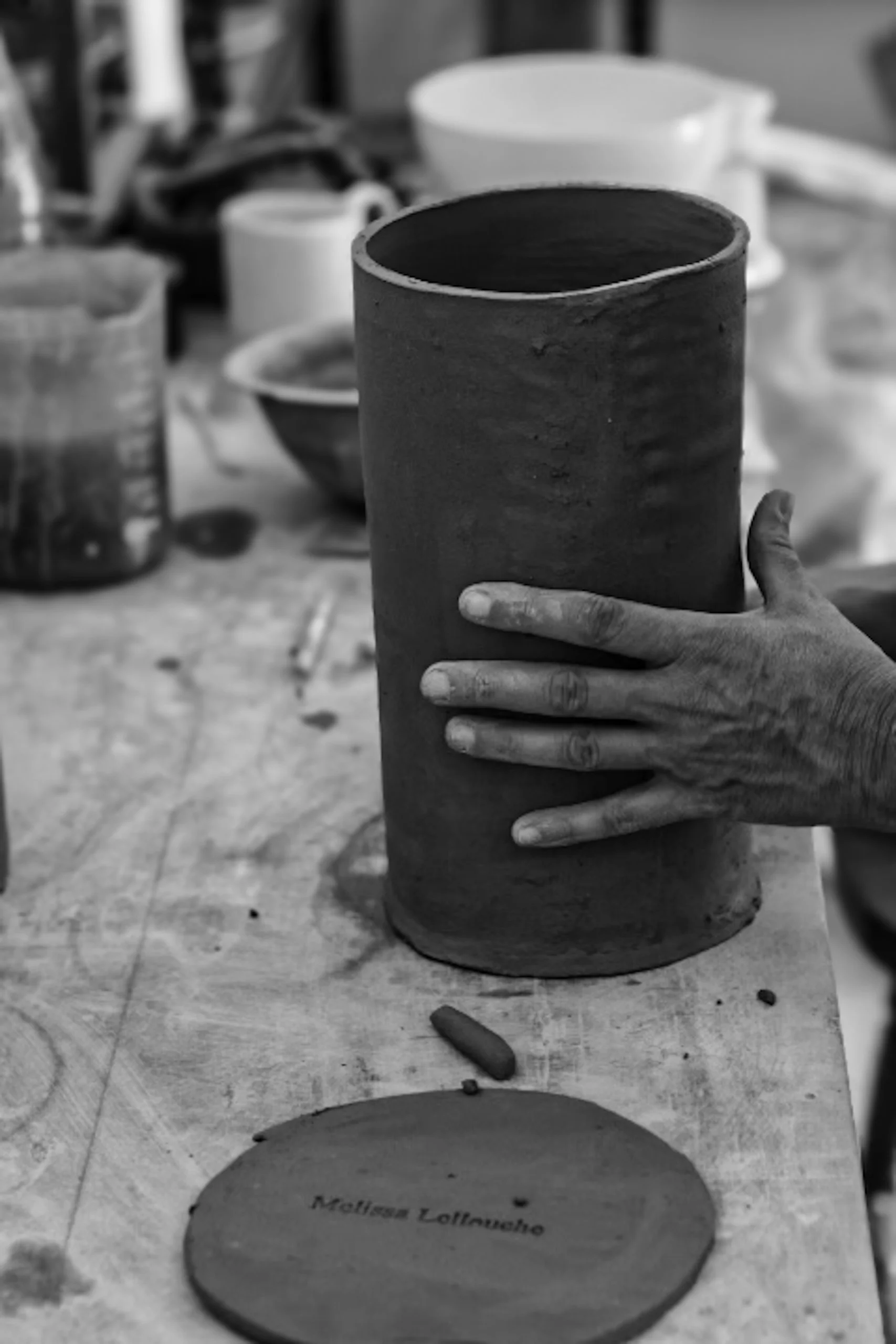 A person's hand holding a tall ceramic pottery piece in a workshop, with various tools and ceramics in the background.