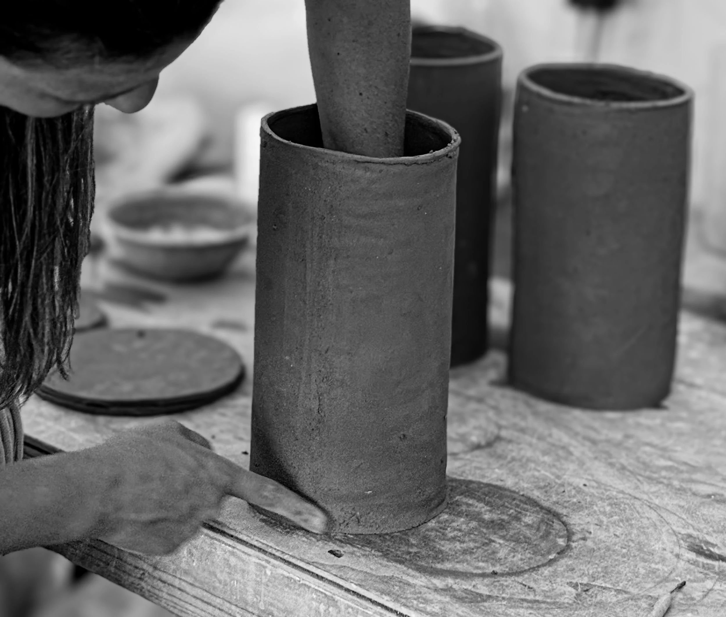 Person working on a clay project, shaping clay inside a cylindrical mold, with other clay molds and tools on a wooden work surface.