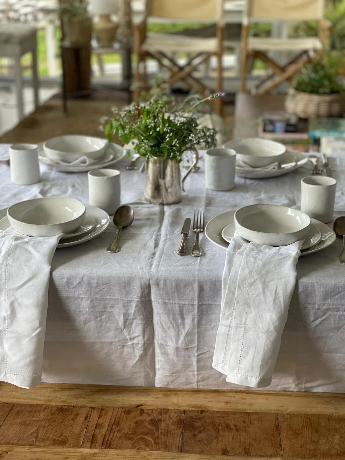 A neatly set dining table with white ceramic plates, bowls, and cups, silverware, and a metal jug filled with a bouquet of small white flowers and greenery, set on a white tablecloth.