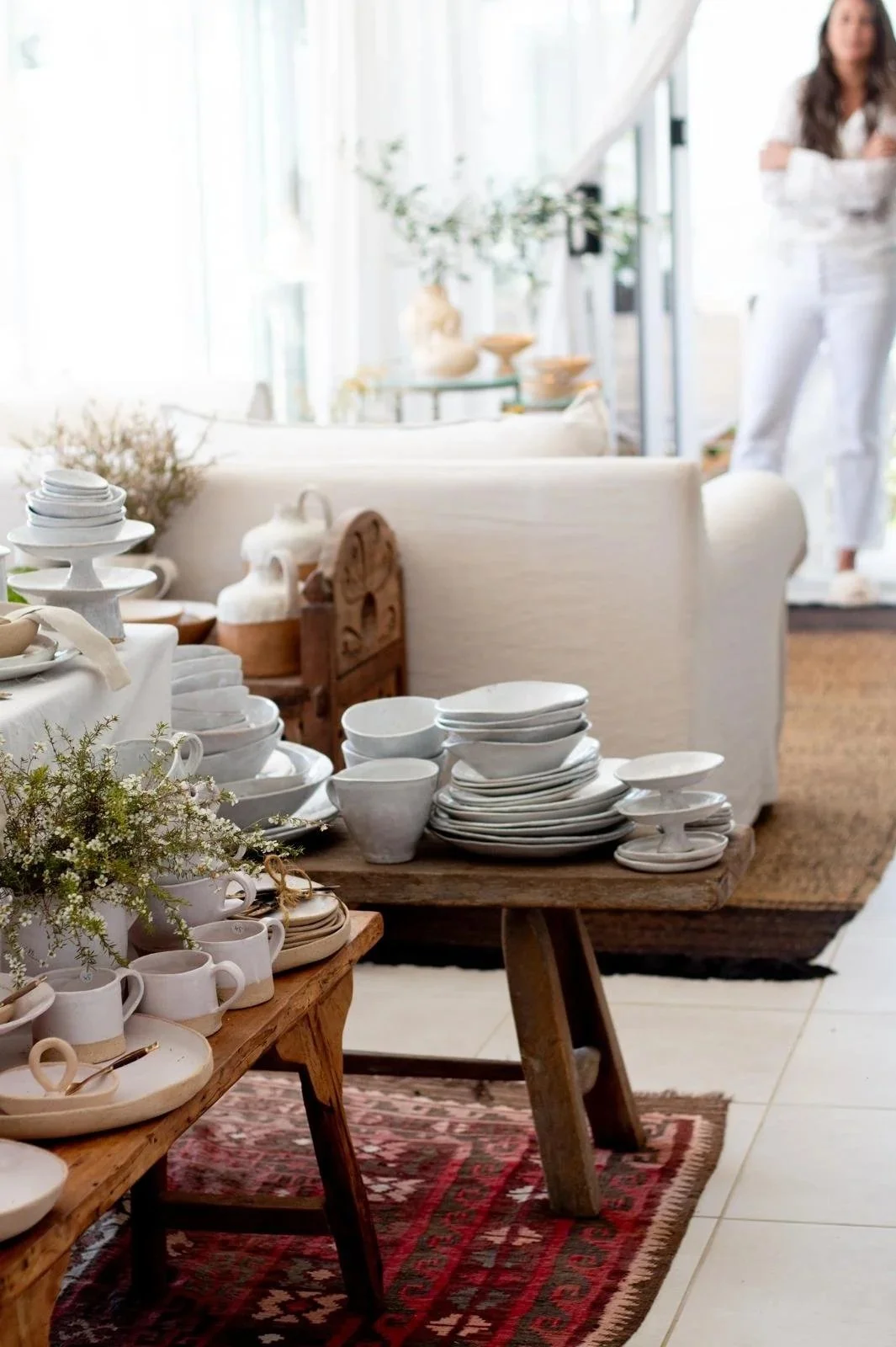 Display of white ceramic tableware and decorative items on wooden tables in a bright, cozy room with a woman standing in the background.