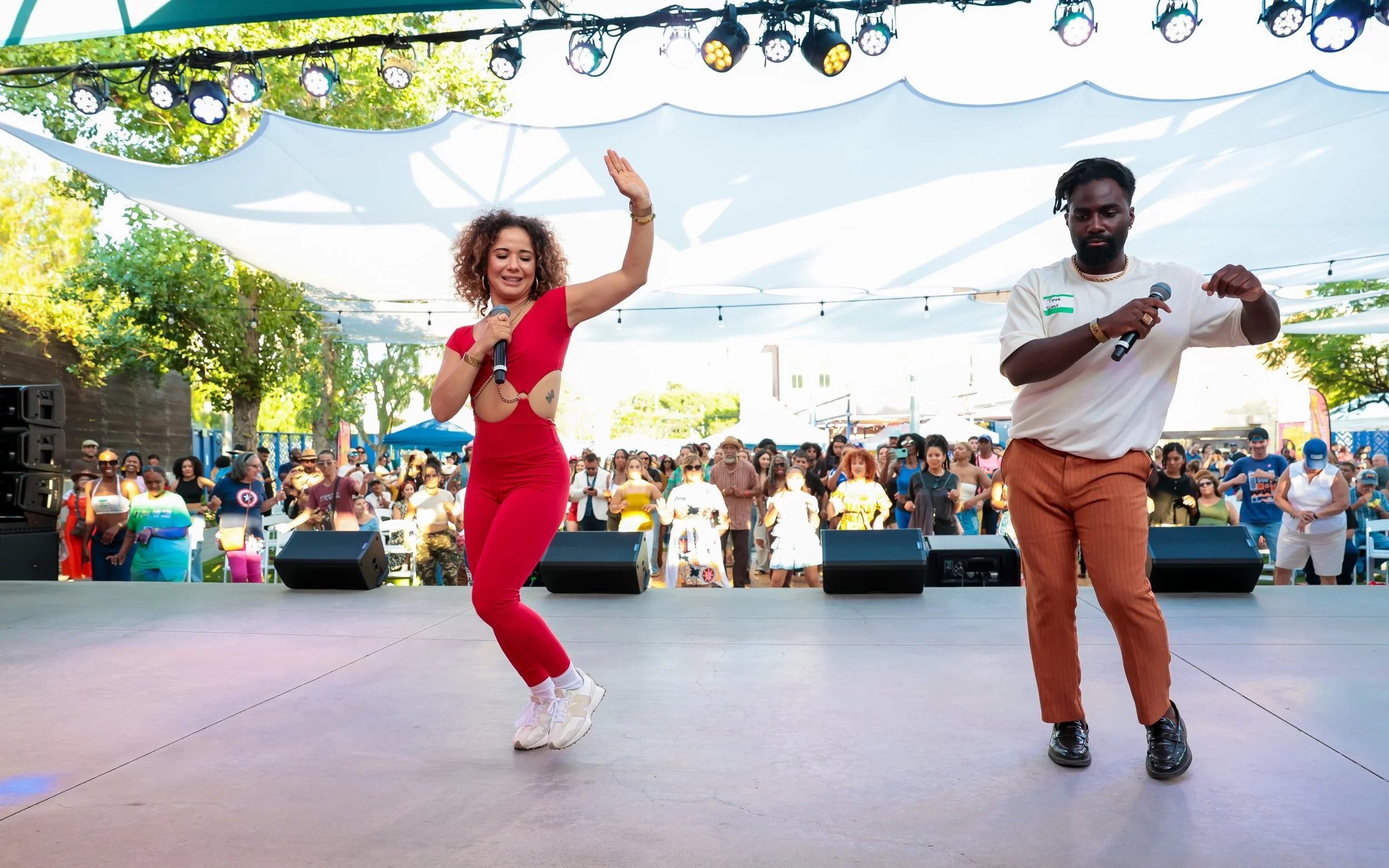 Two performers on stage, one woman in a red jumpsuit and sneakers, dancing with raised hand while holding a microphone, and one man with a beard in a white shirt and brown pants at Afro-Latino Culture Festival.