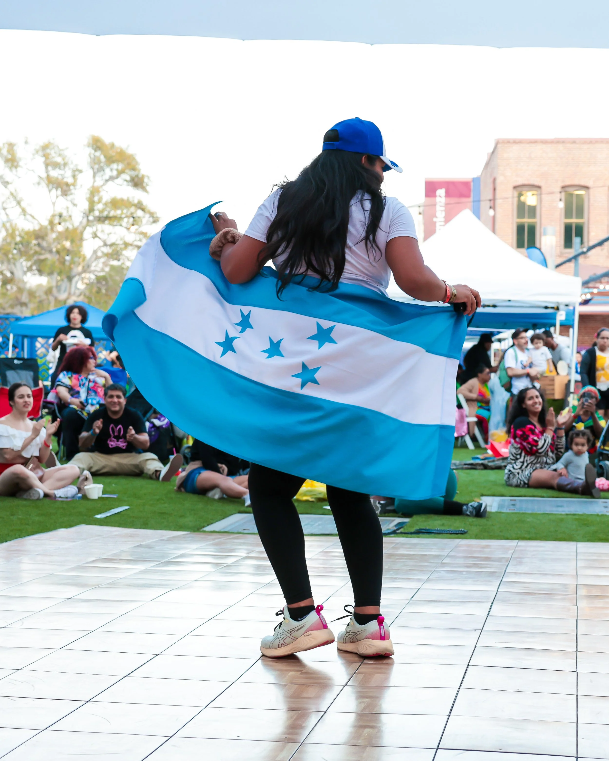 Person in sneakers and black leggings holding a large blue and white Honduras flag, at the Afro-Latino Culture Festival event with seated crowd and tents in the background.