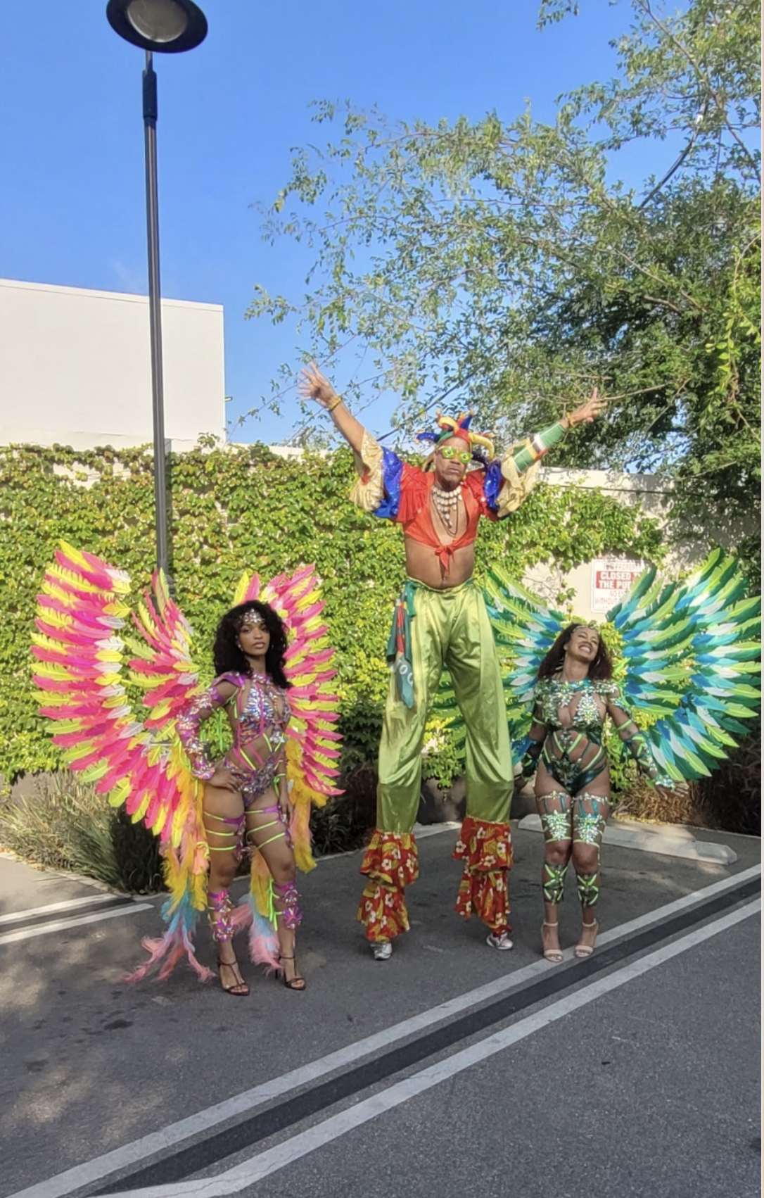 Woman with curly hair, glasses, and hoop earrings smiling at an event. She is wearing a black T-shirt with yellow text that says 'YES! I'SPEAKY SPANISH.' The background features a colorful poster promoting Afro-Latin California.