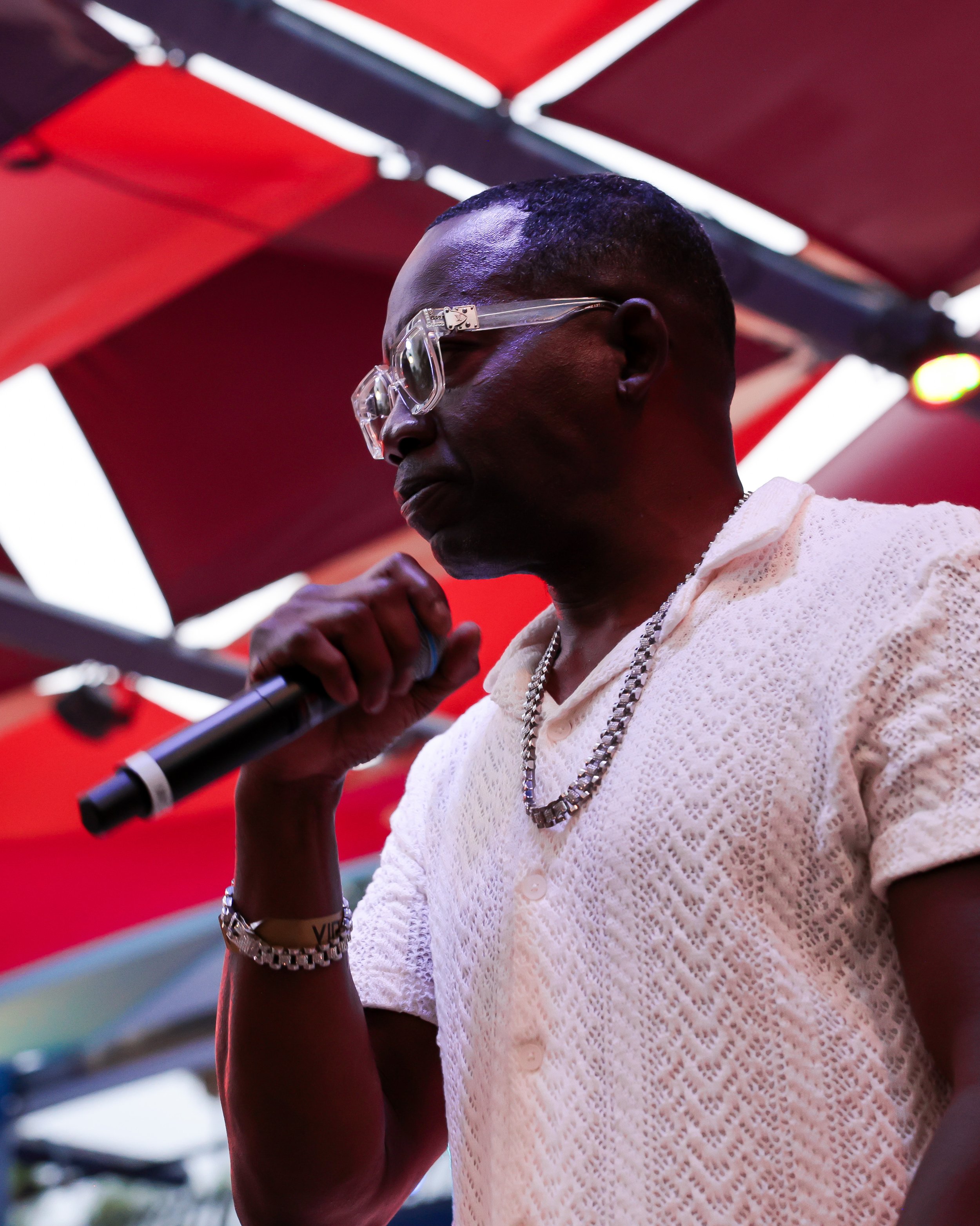 A person with dark skin and short hair, wearing clear glasses, a white textured short-sleeve shirt, a silver chain necklace, and a silver bracelet, standing under a red awning with multicolored lights at the Afro-Latino Culture Festival.
