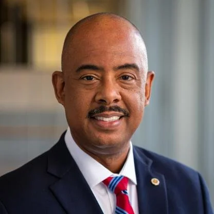 Woman with curly hair, glasses, and hoop earrings smiling at an event. She is wearing a black T-shirt with yellow text that says 'YES! I'SPEAKY SPANISH.' The background features a colorful poster promoting Afro-Latin California.