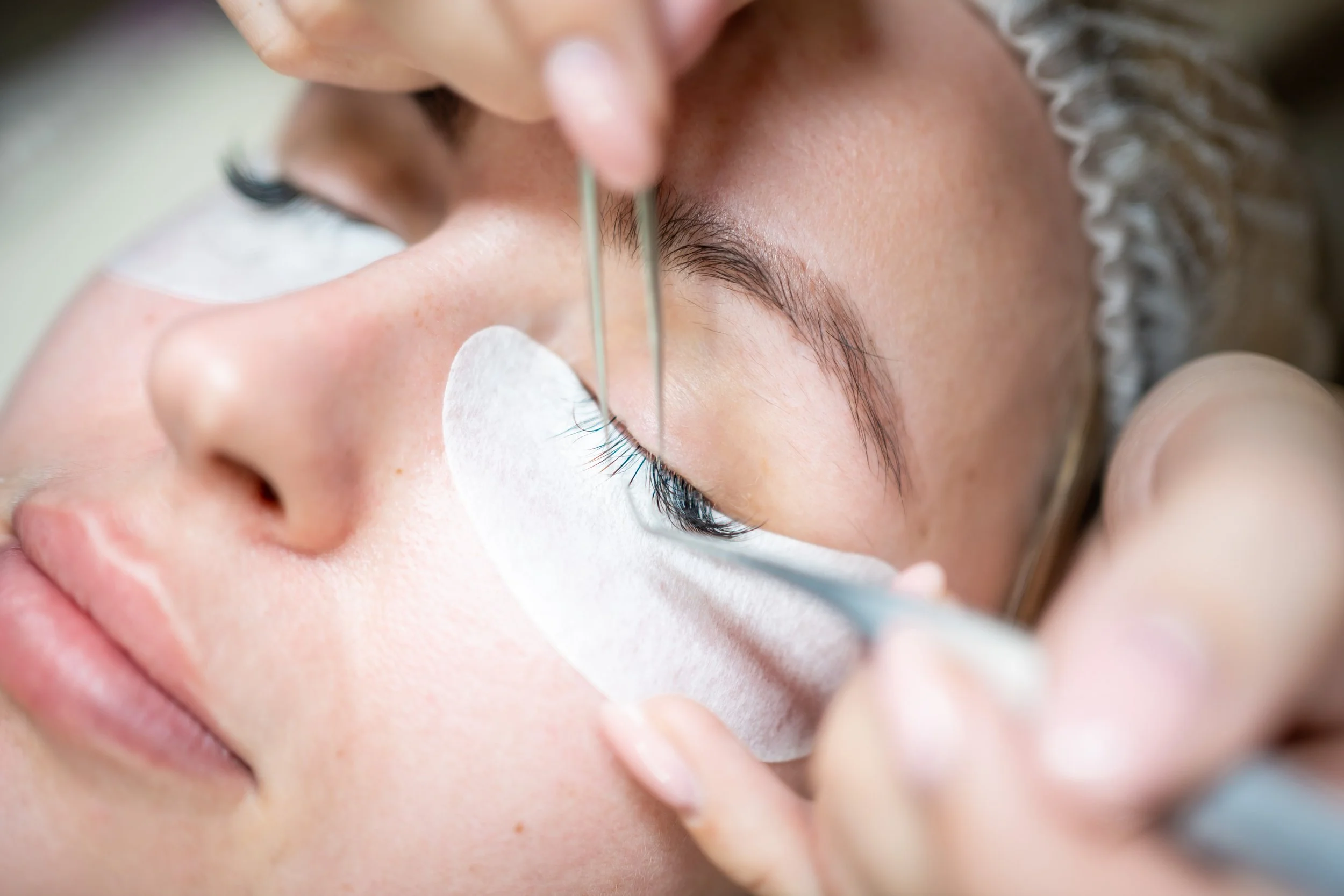 A person receiving a eyelash extension treatment with a beauty technician using tweezers, a protective eye patch and under-eye pads.