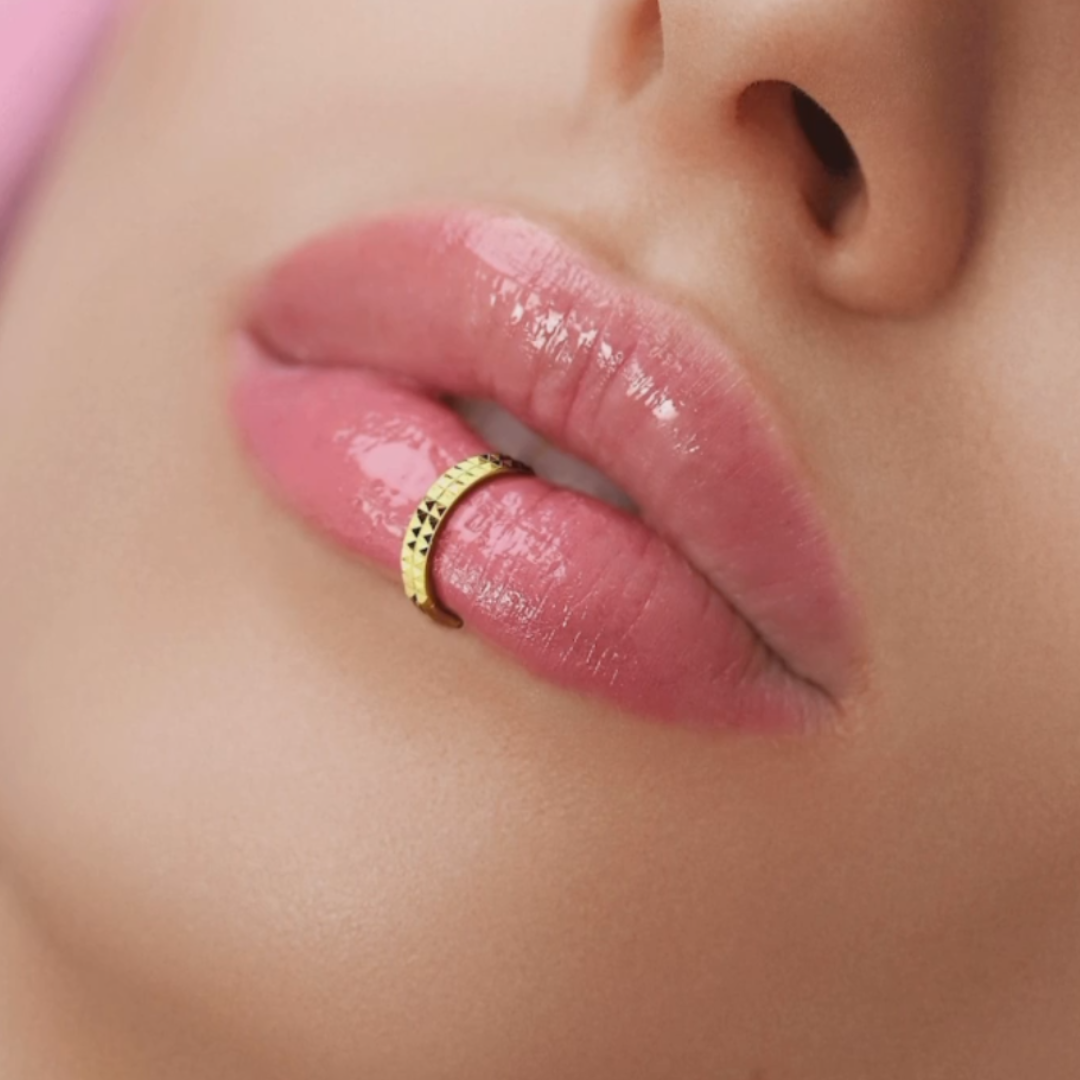 Close-up of a person's lips with pink gloss, holding a gold ring between teeth.
