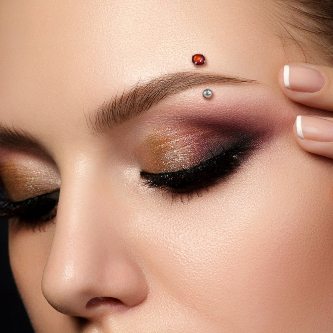 Close-up of a woman's face with eye makeup, a ladybug and a silver ball facial piercing on her forehead, and manicured nails.