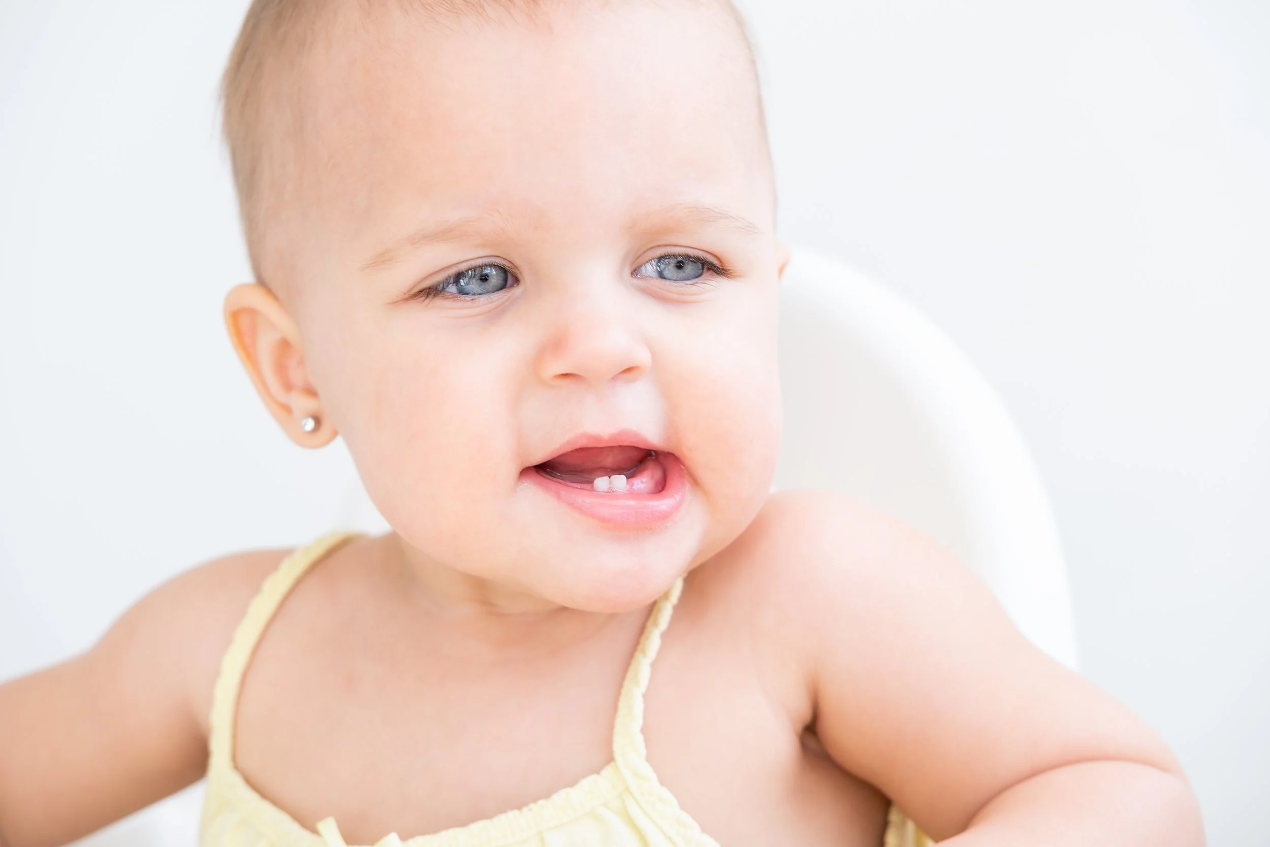 Close-up of a smiling baby with blue eyes, light-colored hair, that just got her baby ear lobe pierced at Salvation Beauty Ink in Pembroke Pines Fl., and wearing a yellow tie-strap outfit, sitting against a plain white background.