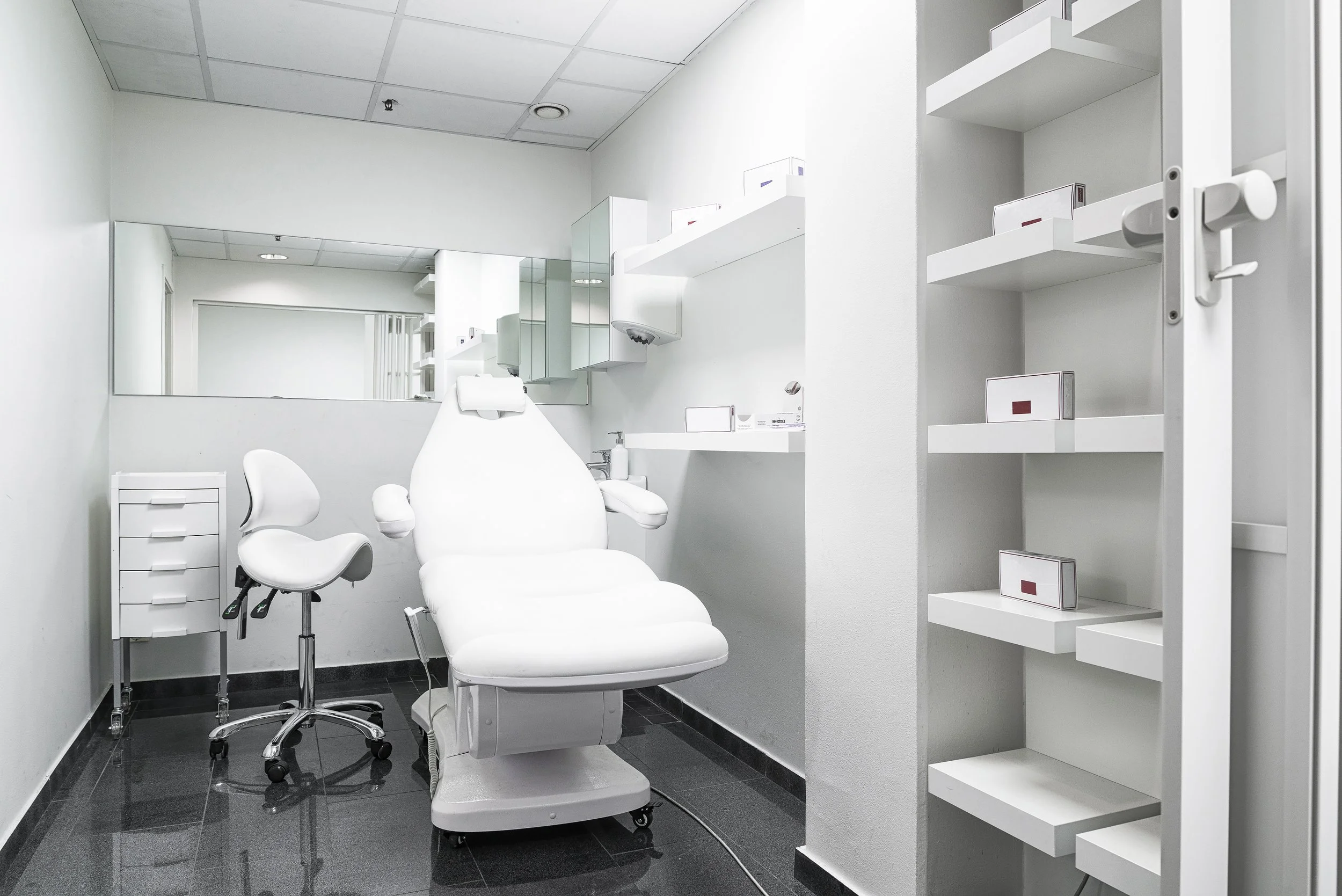 A clean, modern medical or dental examination room with a white examination chair, a white stool, and white shelving units, with a large mirror on the wall.