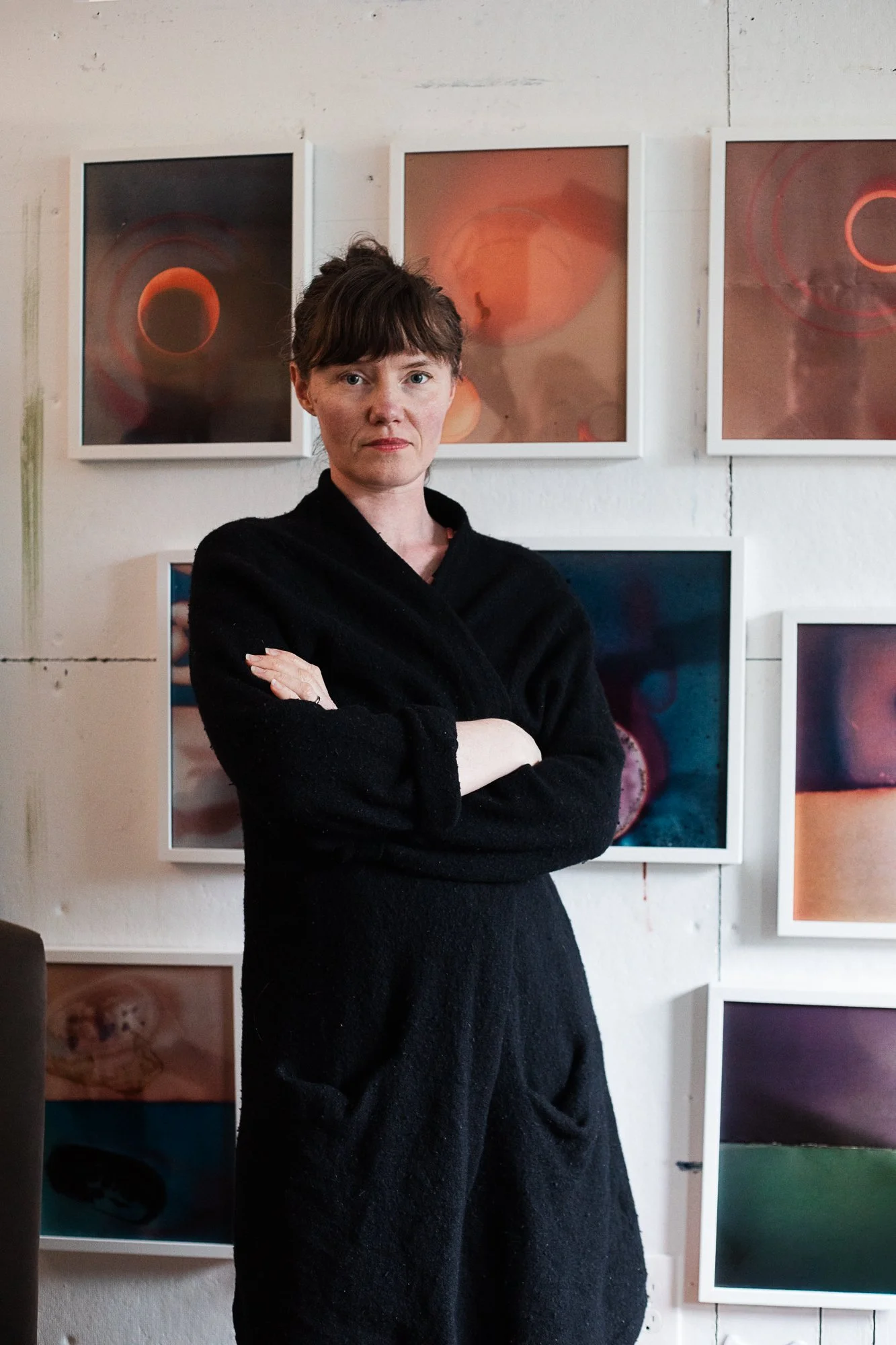 A woman with short brown hair and pale skin standing with arms crossed, wearing a black sweater, in front of a wall with framed artworks featuring abstract circular designs.