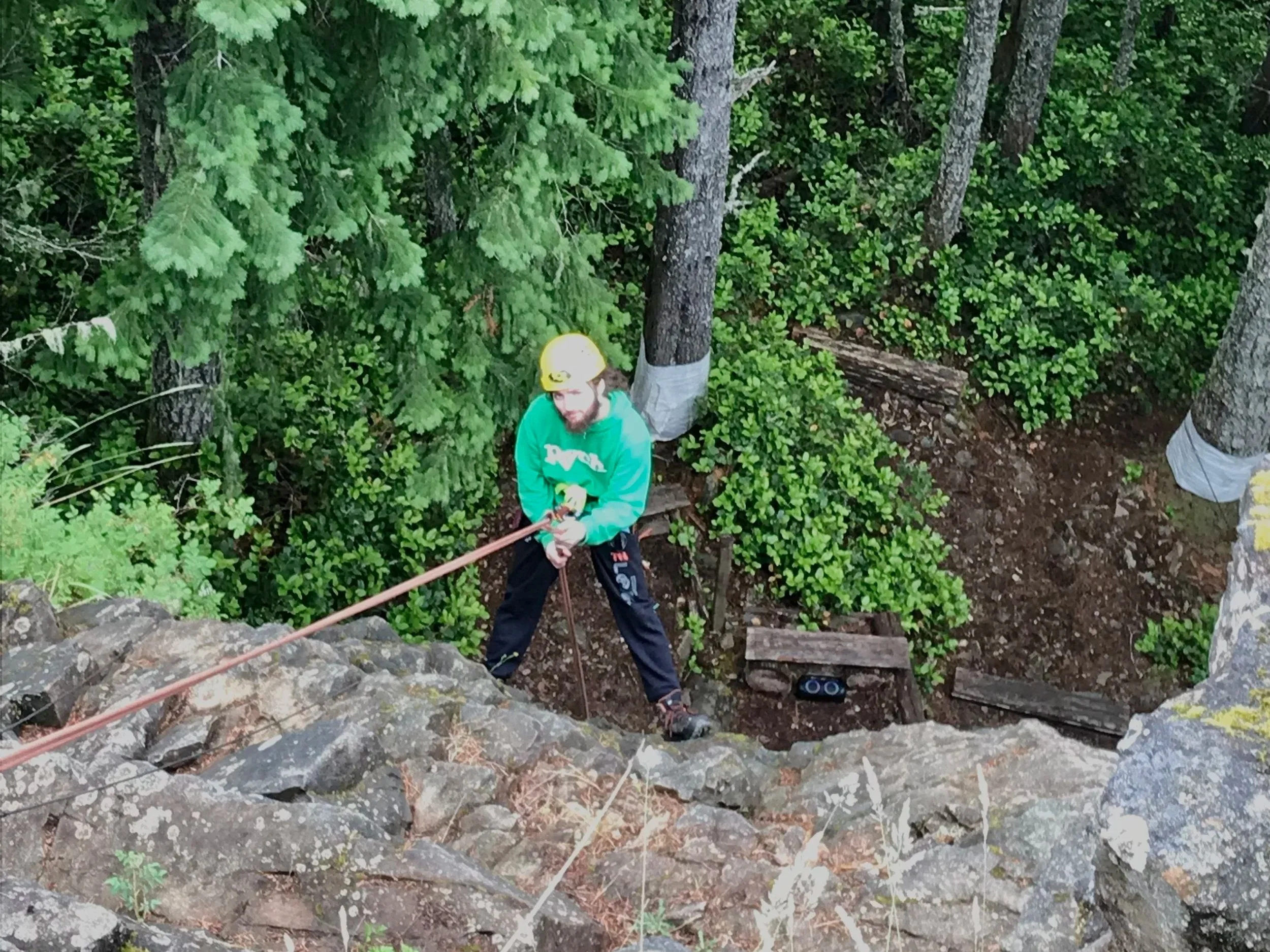 A man wearing a yellow helmet and green hoodie is climbing a rock face using a safety rope in a forested area.