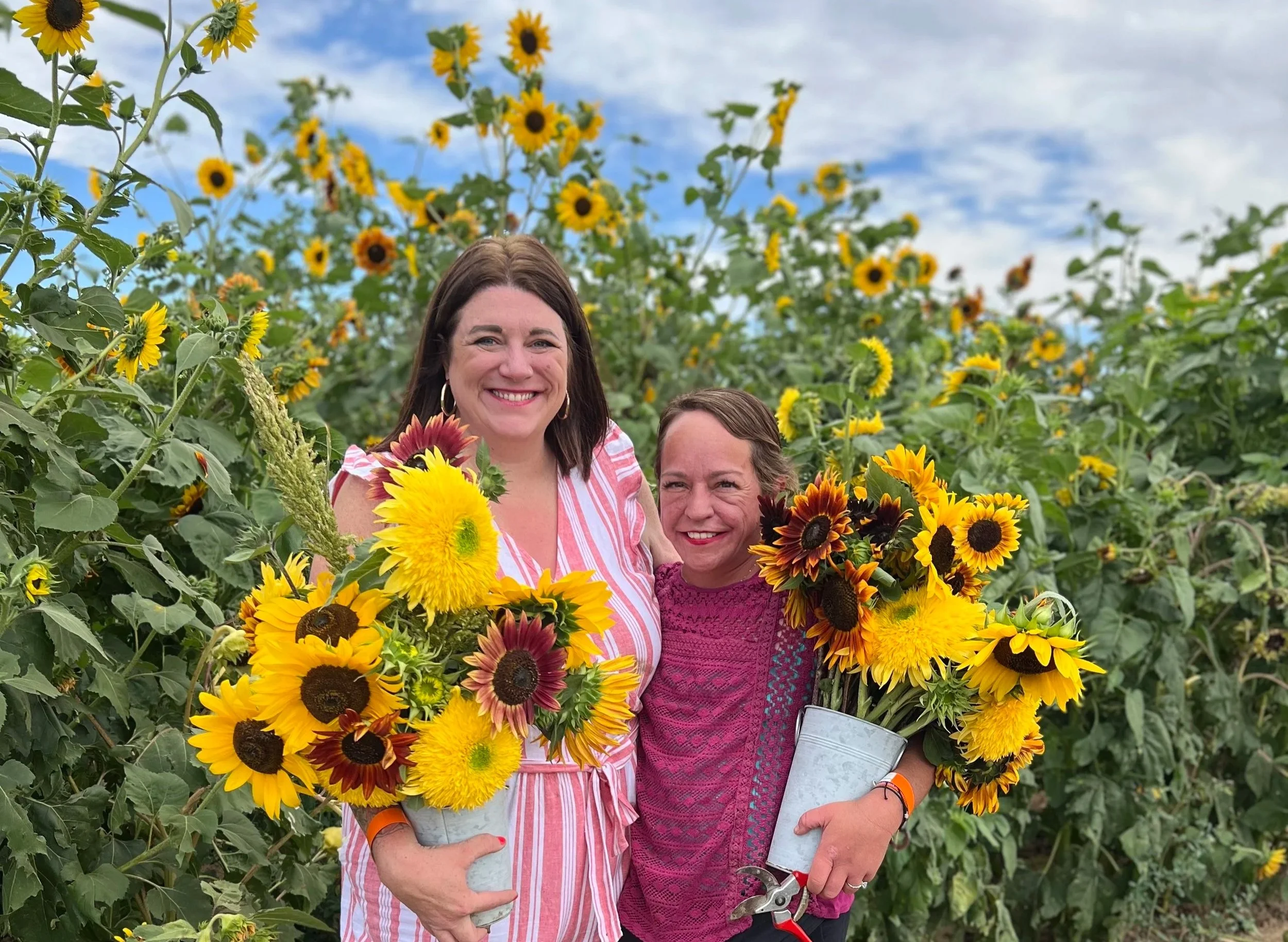 Two women smiling and holding sunflower bouquets in a sunflower field on a partly cloudy day.