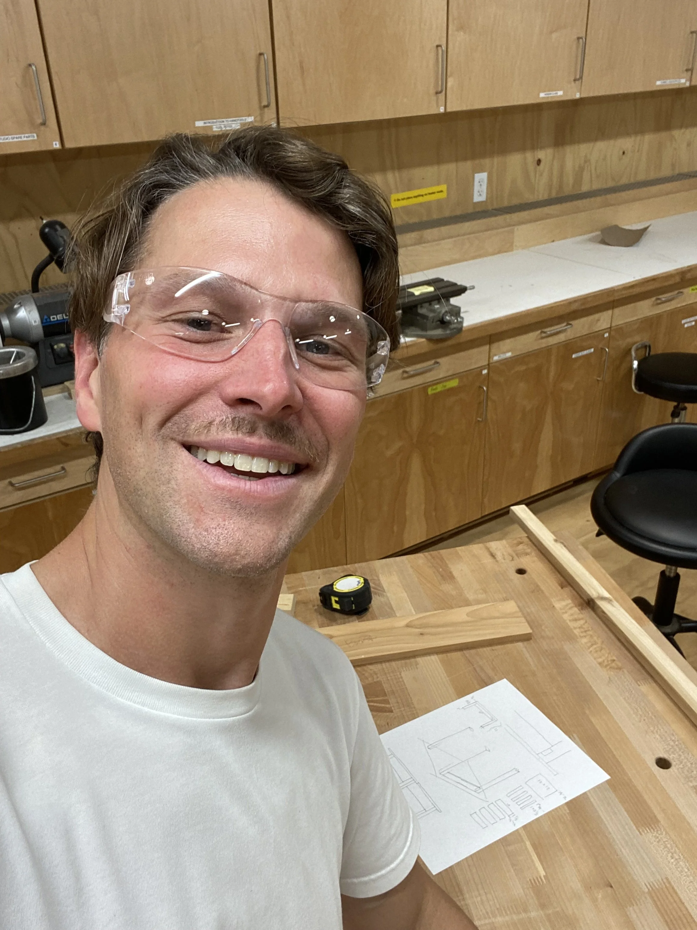 Ben in a woodworking shop, with safety glasses on, a workbench with components and tools visible below him