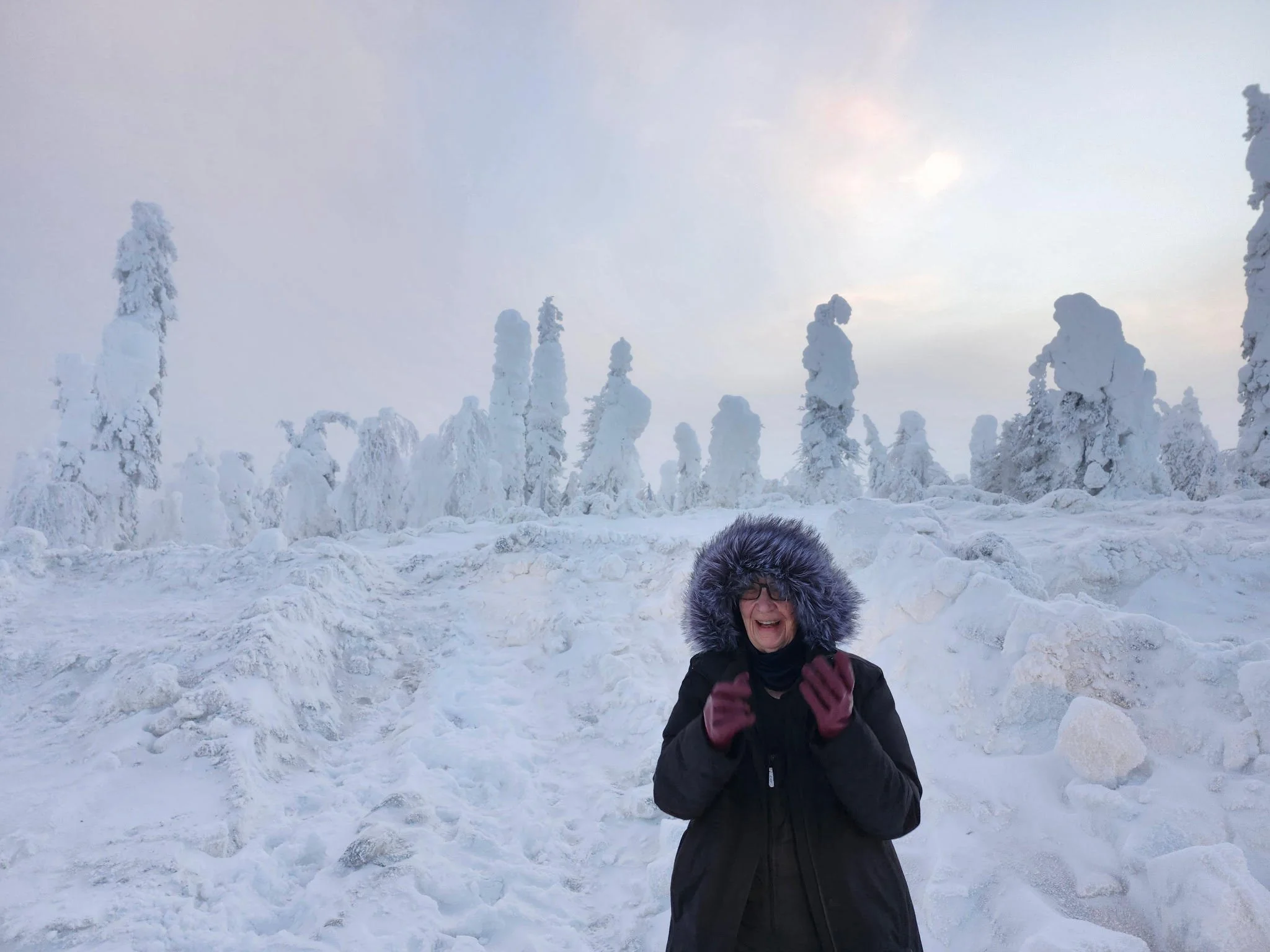 Jane is wrapped in a giant black parka with a massive fur edged hood, big red gloves, and she is laughing. In the background is snow everywhere. Trees piled high with mounds of snow. She is in Alaska close to the Arctic Circle.