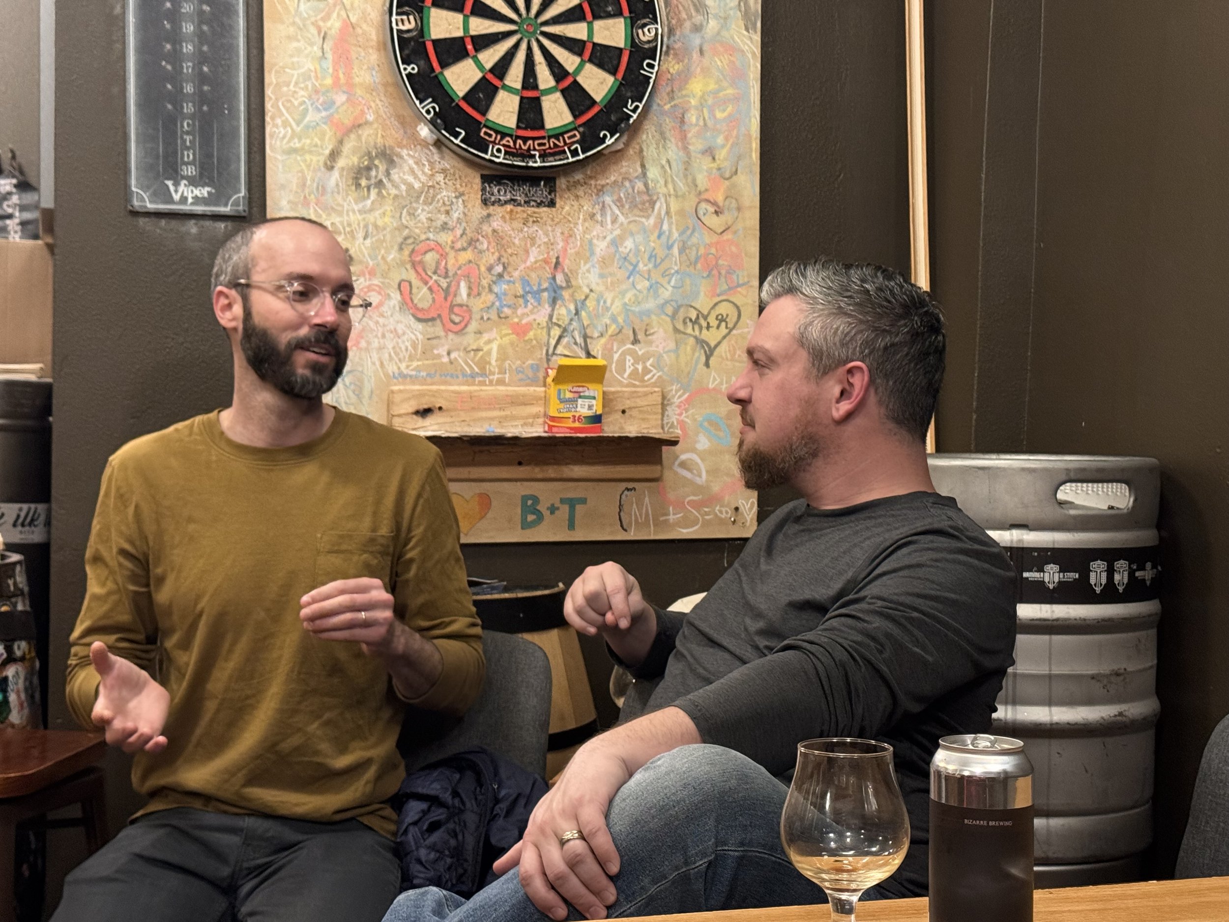 Andrew in a mustard shirt, wire-rimmed glasses, and hands expressing his ideas chatting with Ryan, wearand Ryan wearing jeans a dark grey long sleeved shirt, legs casually crossed listening to Andrew.