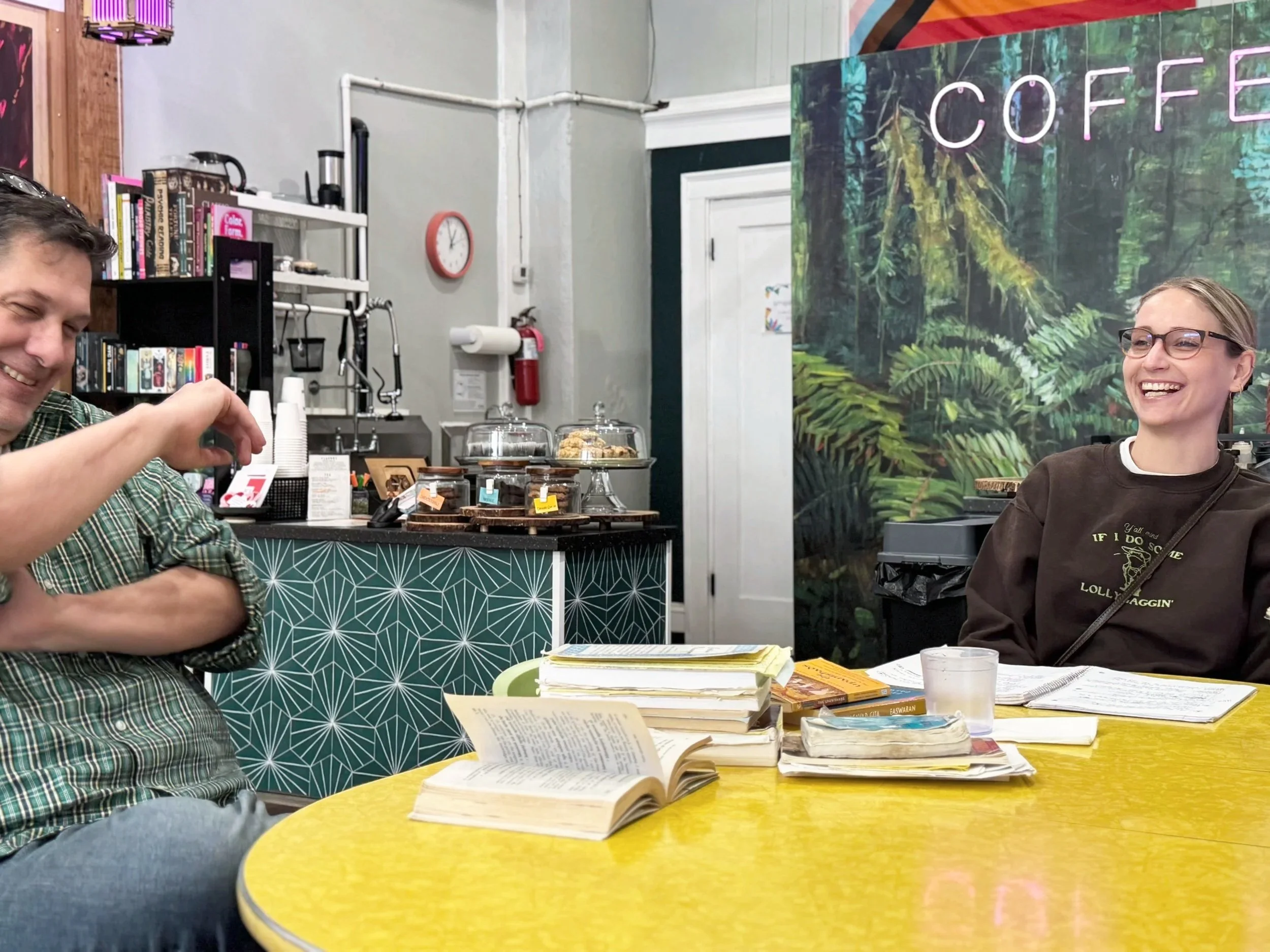 Ciera and Seth laughing during their tutorial studies of South asian thought. They sit at a yellow table stacked with books and notes at Cambium Coffee & Gallery