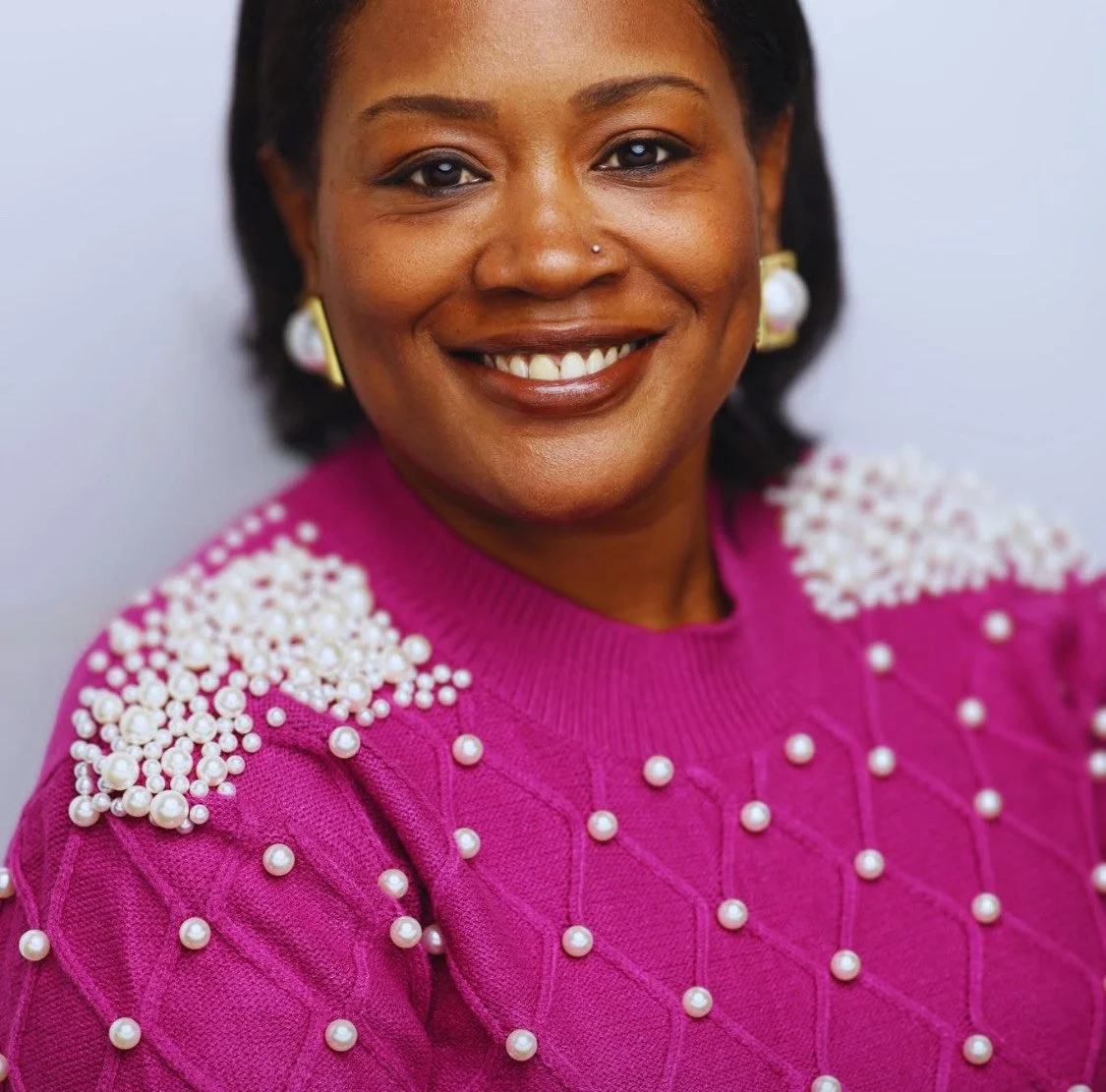 Smiling person wearing a pearl-embellished magenta sweater and pearl earrings.