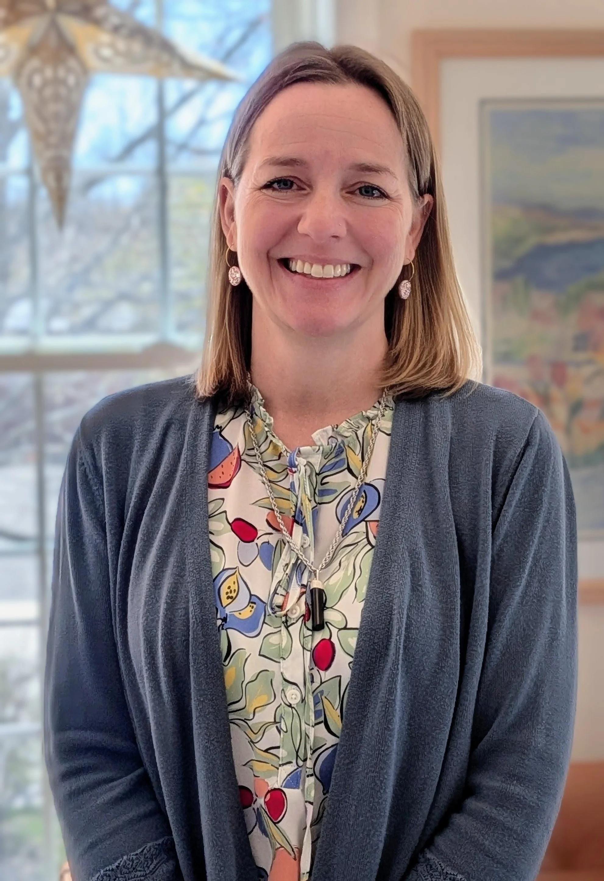 A photograph of Melissa Tarbox, a woman with shoulder-length blonde hair smiling, wearing a floral blouse, earrings, and a grey cardigan, indoors with a window and artwork in the background.