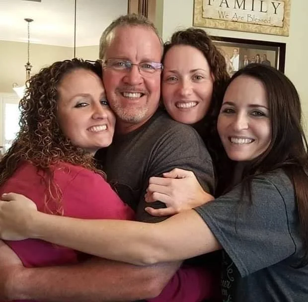Four people hugging and smiling in a family photo, with a sign in the background that reads "Family, We Are Blessed."