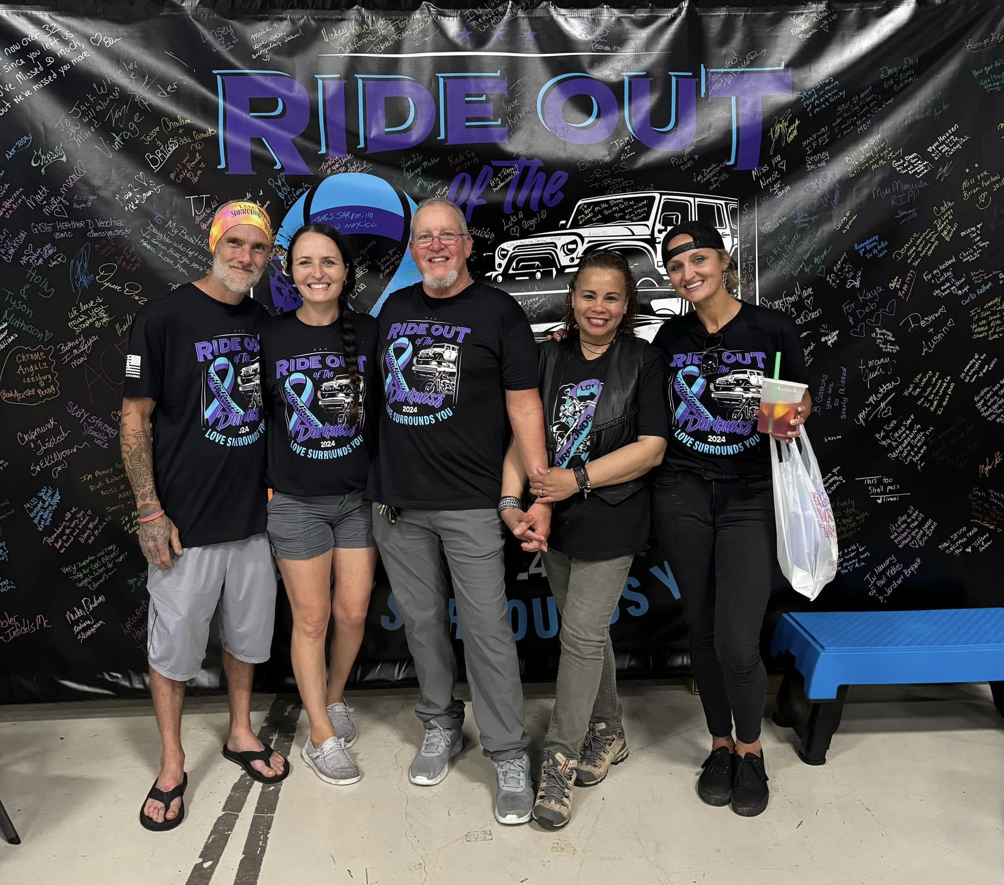Group of five people standing together in front of a black backdrop with handwritten messages, all wearing matching black T-shirts with a design that says 'Ride Out of the Darkness 2024' and an illustration of a vehicle with a purple awareness ribbon. The group is smiling, with one person holding a drink and a bag, and the backdrop features a white outline of a vehicle and colorful signatures.