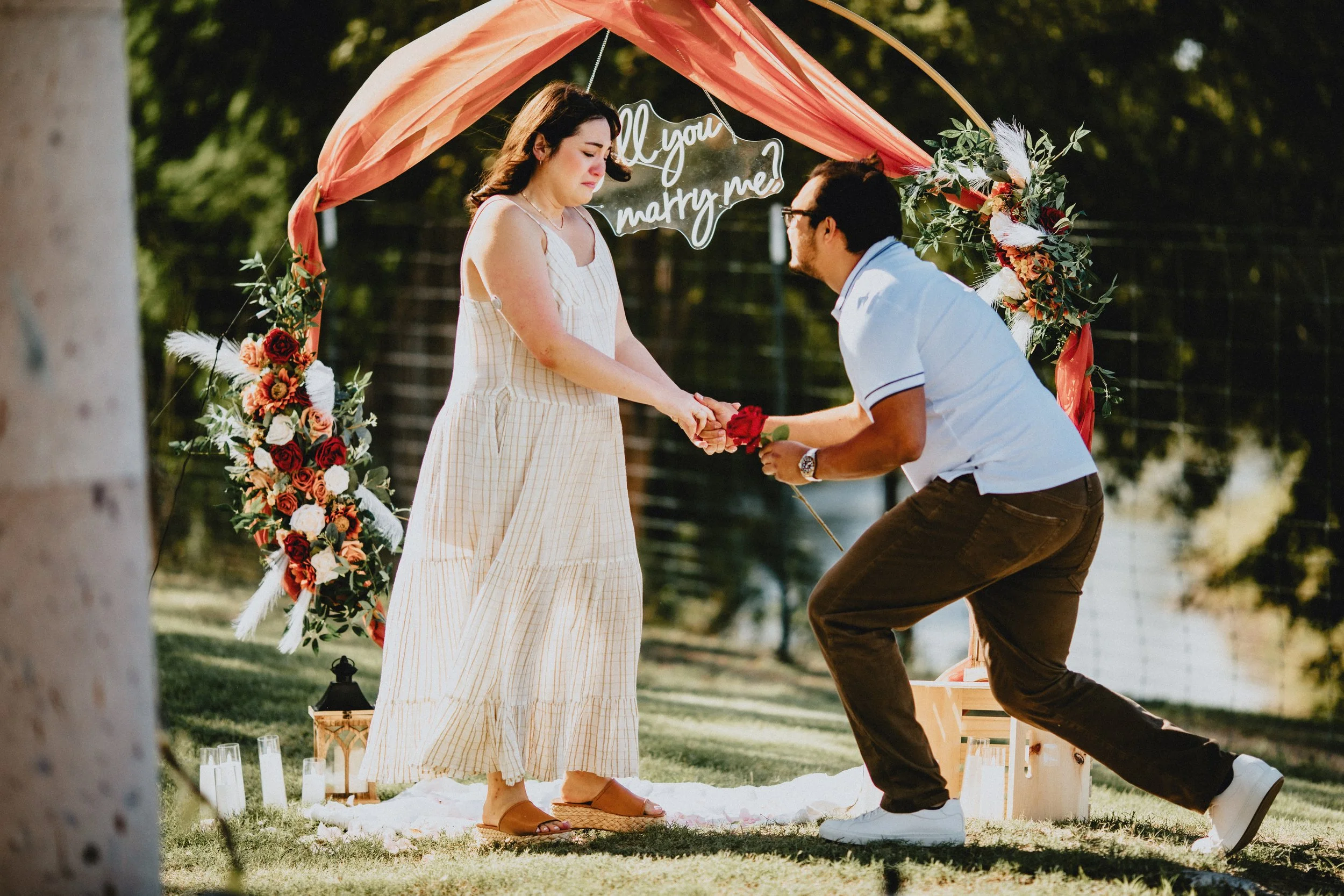 Man proposes to woman during outdoor wedding ceremony at sunset, with a floral arch and a sign that says 'Will you marry me?' in the background.