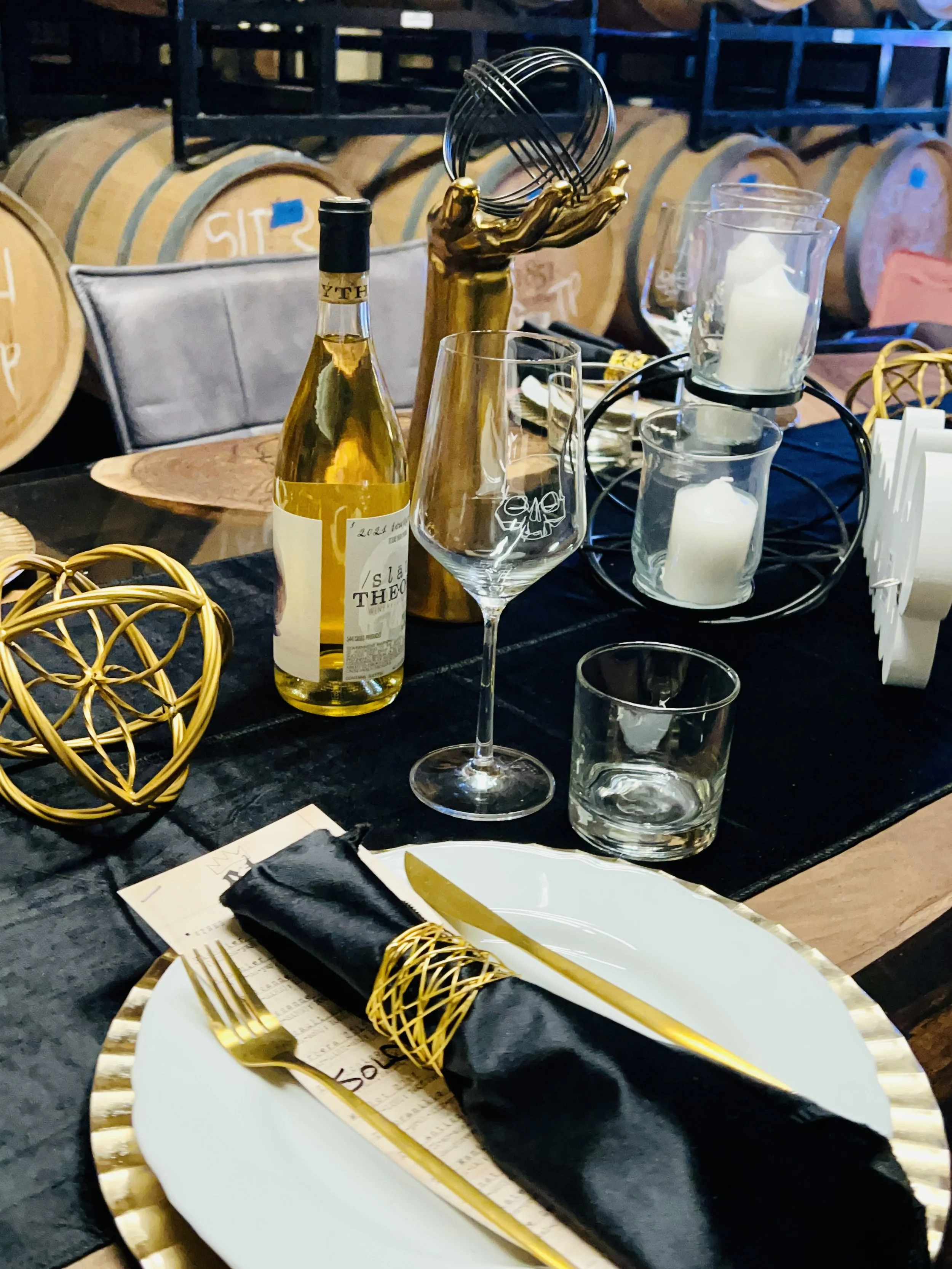 A table setting featuring a white plate with a black napkin wrapped around gold utensils, a gold decorative sphere, a wine glass, a water glass, and a bottle of white wine. There are candles and decorative items on the table. In the background, there are wine barrels and shelves.