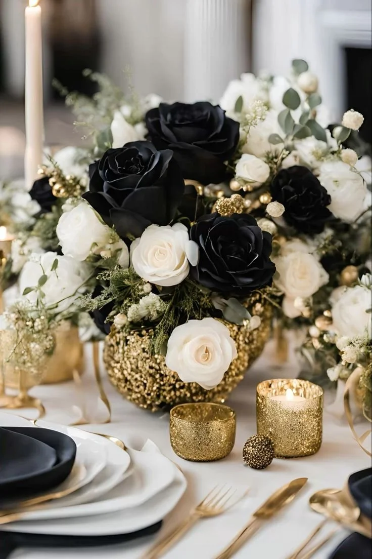 Elegant table centerpiece with black and white roses, gold accents, candles, and tableware for a formal event or celebration.