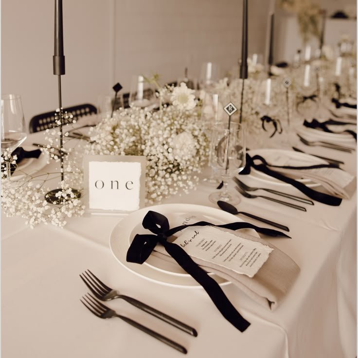 Elegant wedding reception table decorated with white flowers, black napkins, and gold accents, with a small sign reading 'one' and a menu card tied with black ribbon.