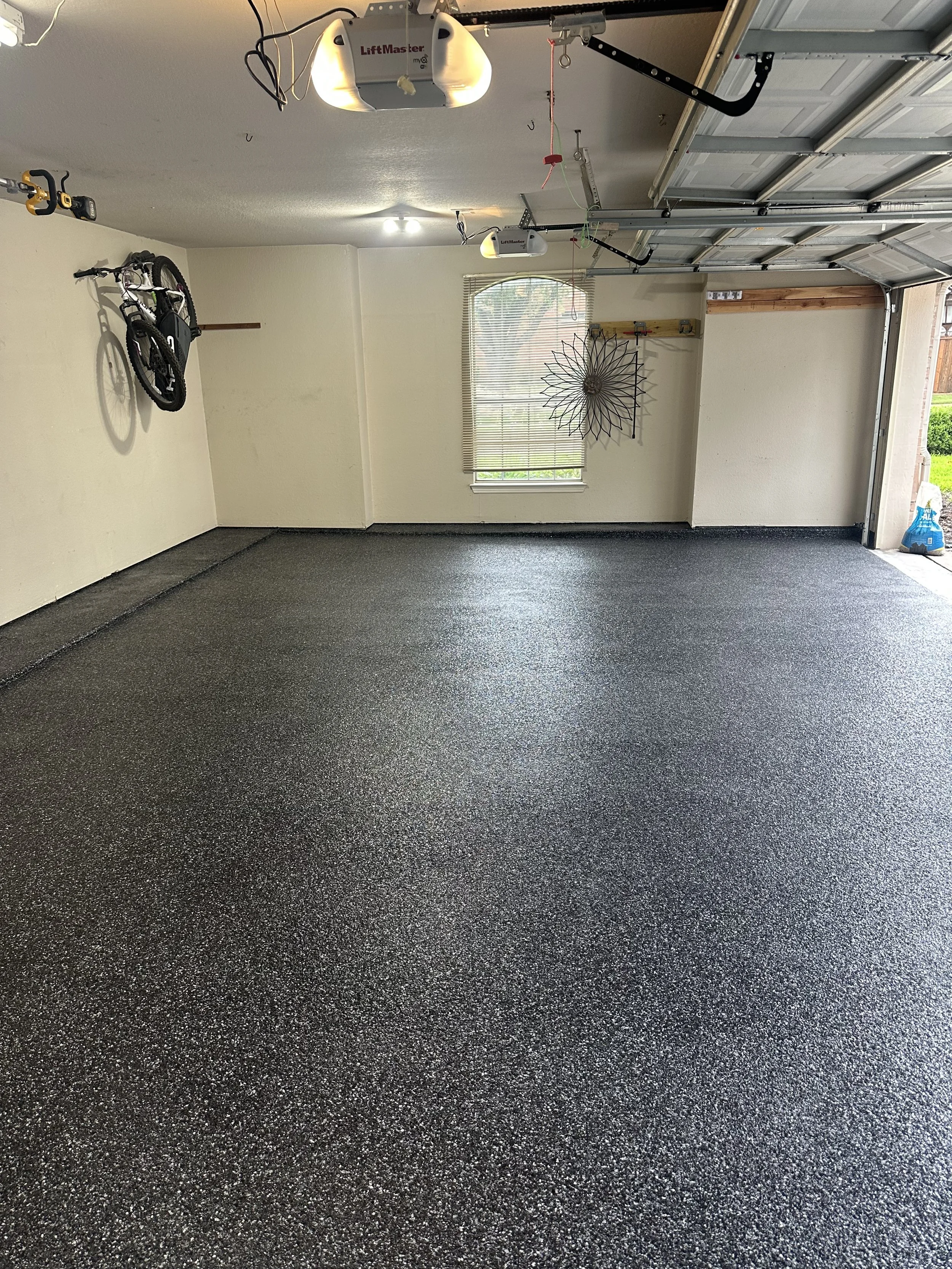 Empty garage with a mounted bicycle on the wall, a window with blinds, a wall decoration, and a garage door with an automatic opener. The floor is covered with a carbon black speckled epoxy coating.