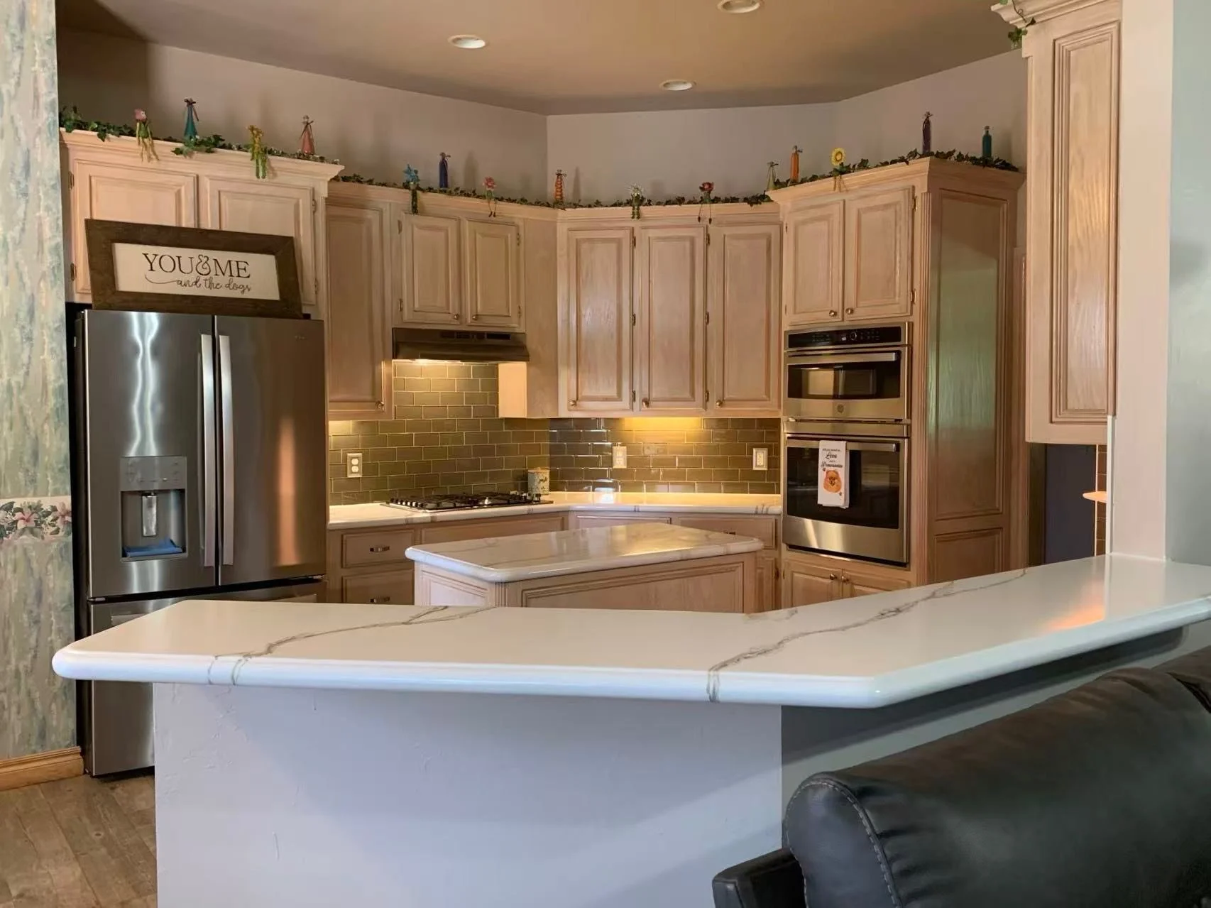 Kitchen with light wood cabinets, stainless steel refrigerator and appliances, brown epoxy backsplash, white epoxy marble countertop, and decorative figurines on the upper cabinets. A white epoxy marble island in the foreground.