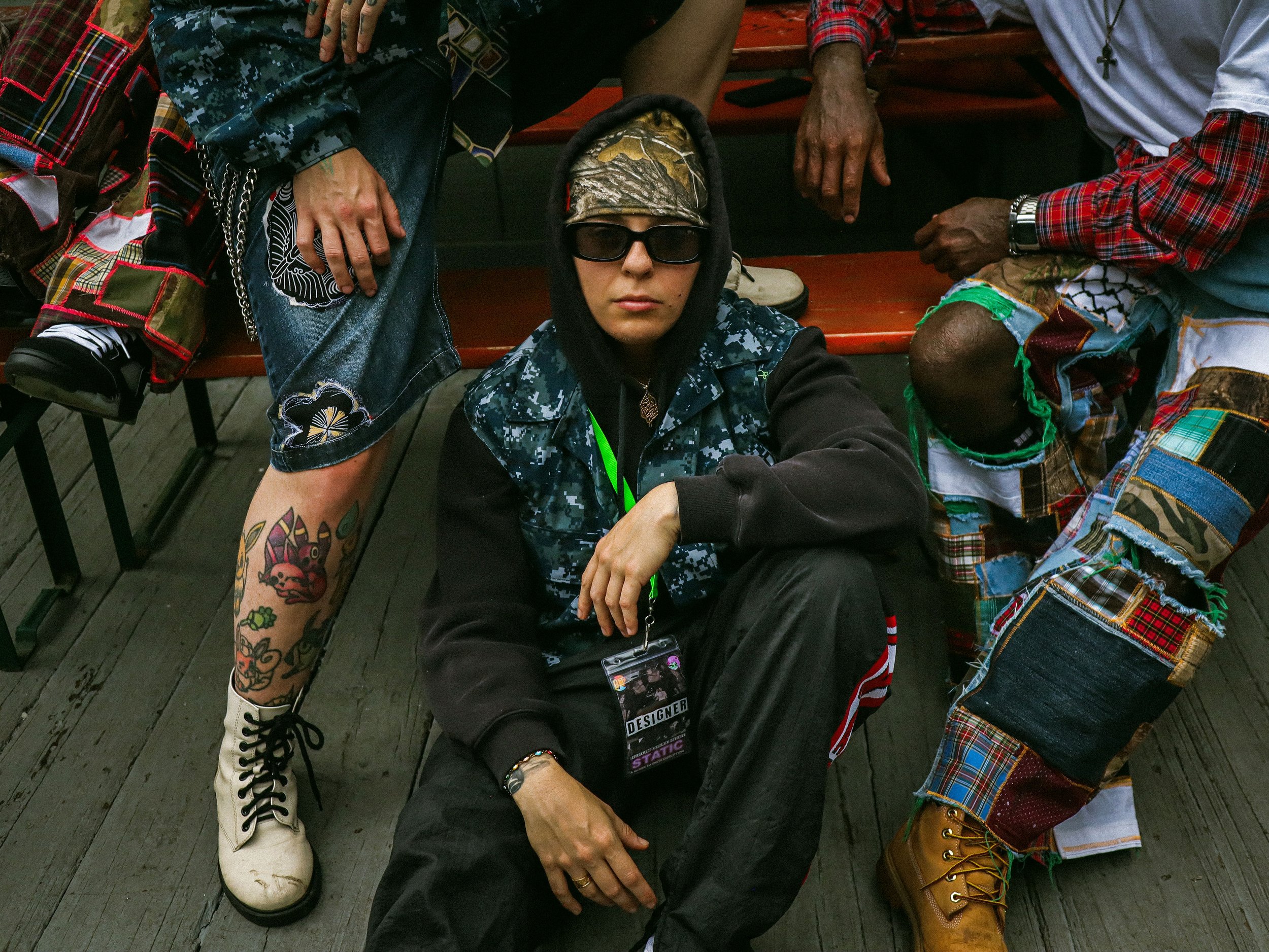 A fashion designer sitting on the ground surrounded by models, with tattoos, a hoodie, and a military-style jacket featuring patches, dressed in colorful, patterned handmade clothing, some with plaid patterns, and boots.