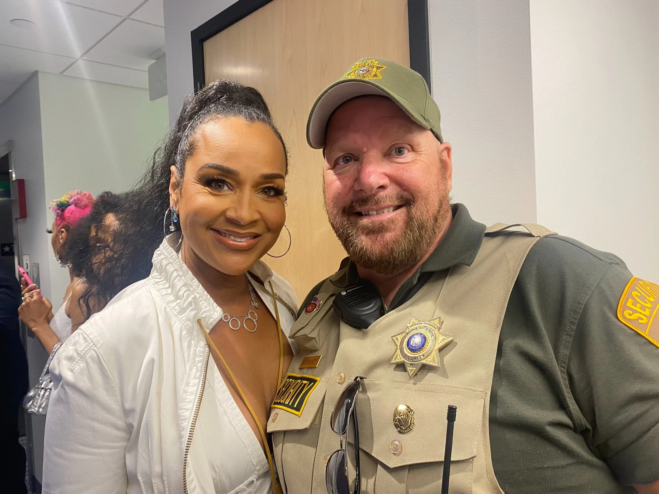 A woman and a security officer smiling and posing for a photo indoors. The woman is wearing a white jacket and jewelry, and the officer is in uniform with a badge and patches.