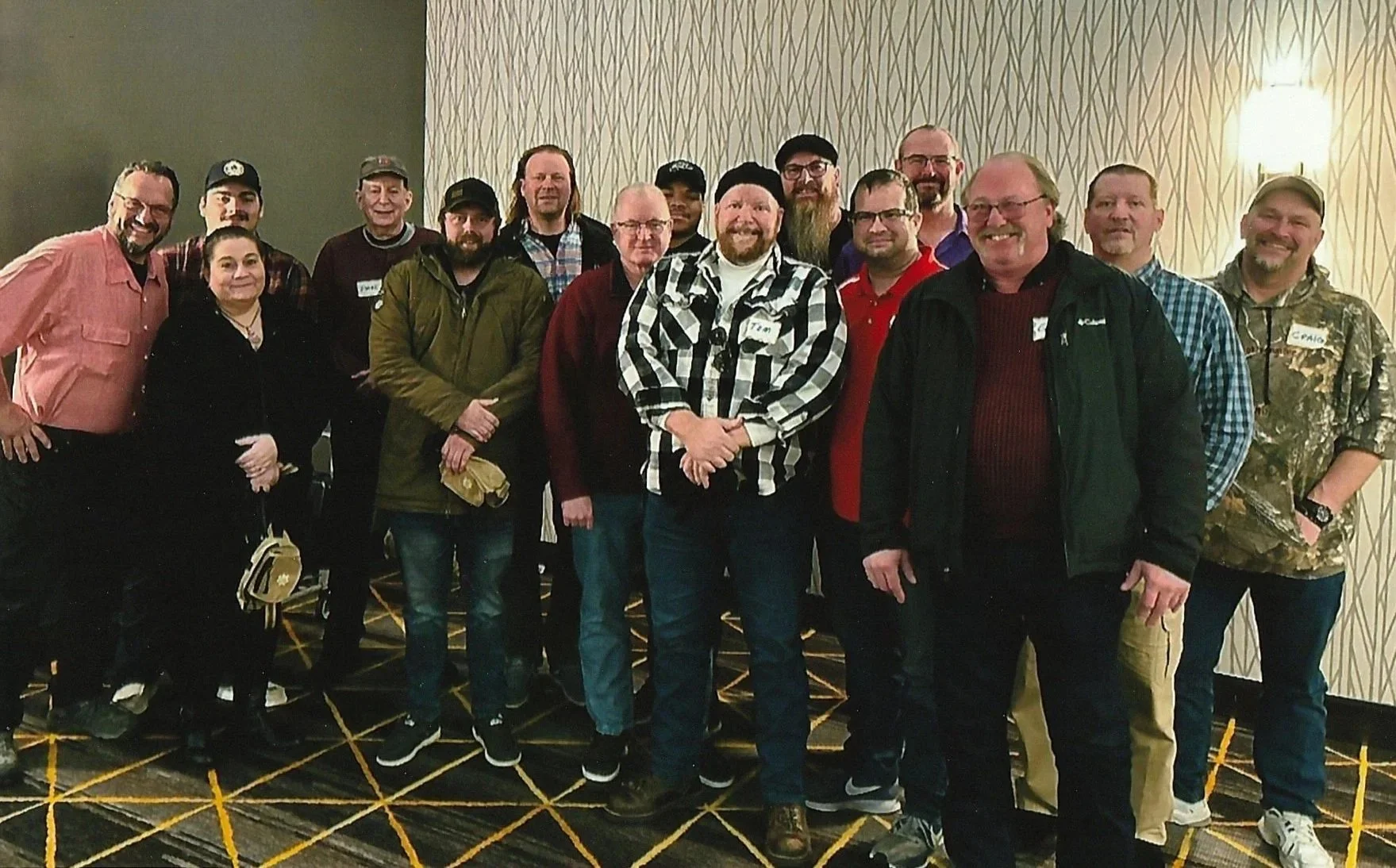 Group of fifteen people, mostly men, posing together in an indoor setting with patterned carpet and wallpapered walls, some wearing name tags.