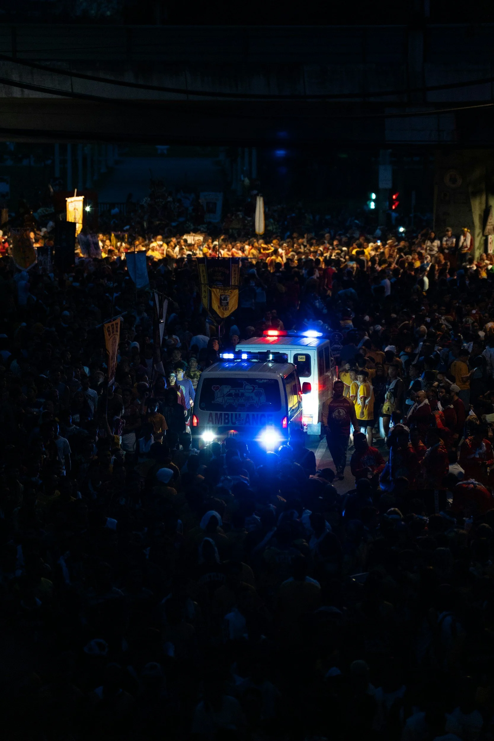 A large crowd fills a dark area with an ambulance parked in the center, illuminated by bright blue and red emergency lights.